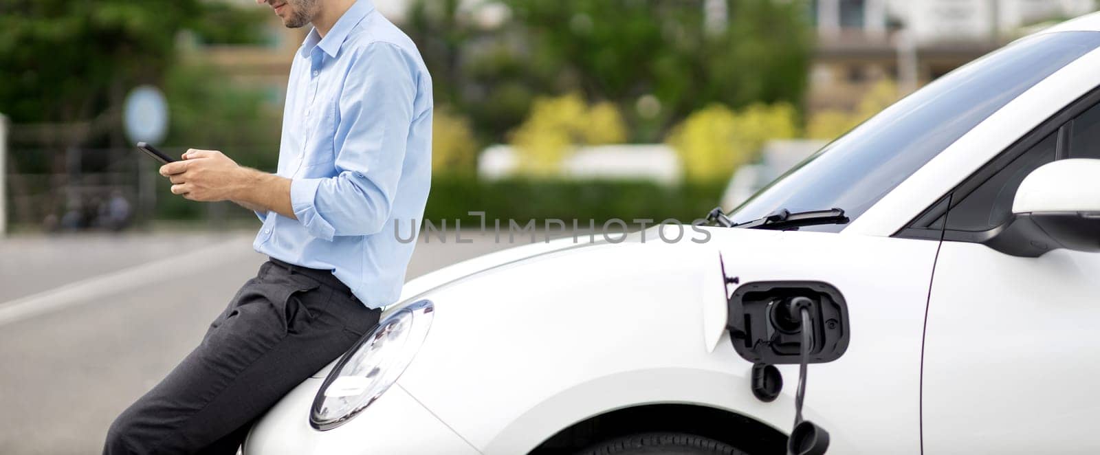 Closeup progressive suit-clad businessman with his electric vehicle recharge his car on public charging station in modern city with power cable plug and renewable energy-powered electric vehicle.