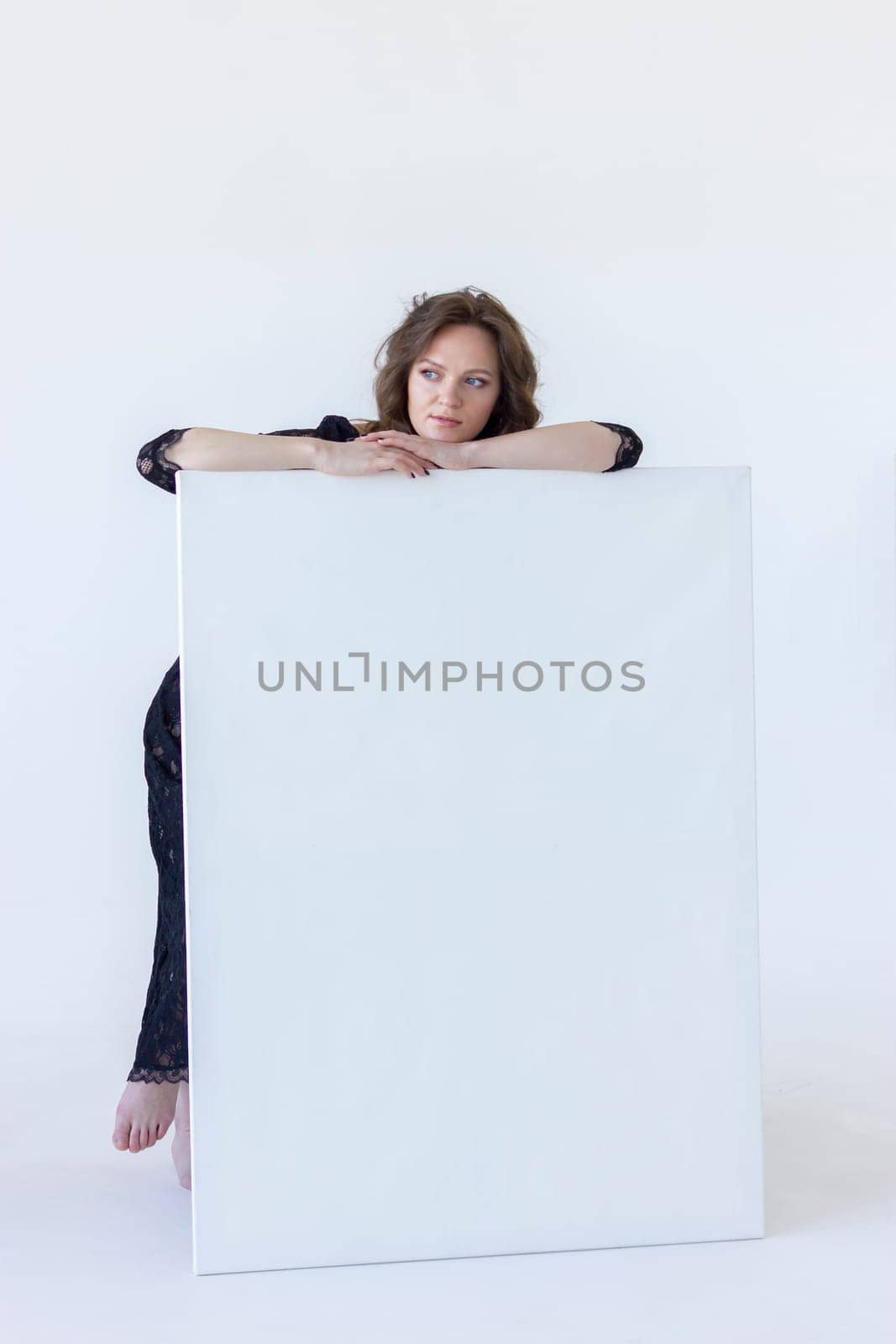 Young Woman Leans On White Blank Large Stretched Canvas for Painting. Beautiful Female Artist Poses With Empty Boards On White Background Indoor. Mockup. Vertical Plane. High quality photo