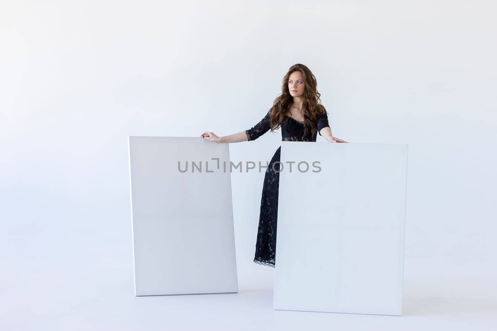 Young Woman Stands Between White Blank Large Stretched Canvases for Painting. Beautiful Female Artist Poses With Empty Boards On White Background Indoor. Mockup. Horizontal Plane. Copy Space by netatsi
