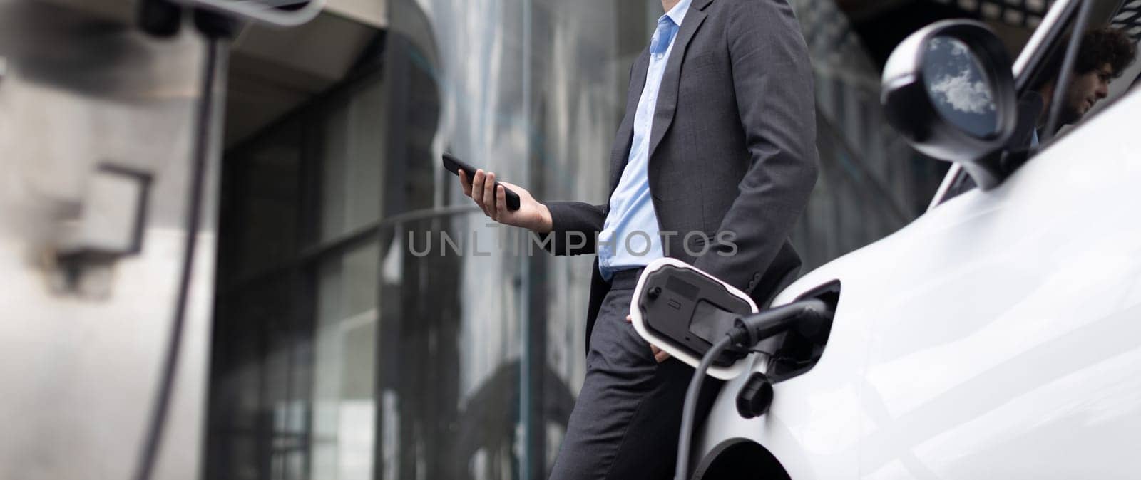 Closeup progressive businessman with electric car recharging at public charging station with background of city residential building. Eco friendly car powered by alternative clean energy.