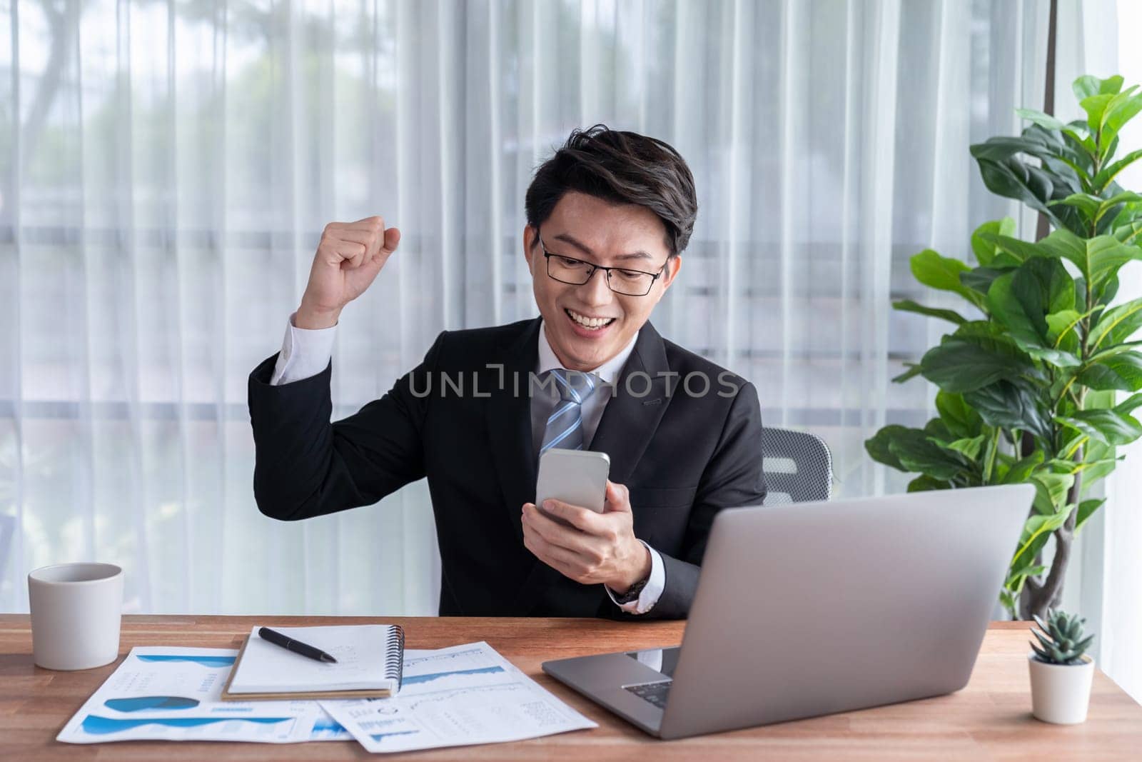 Excited Asian businessman celebrates success at office desk. Happy office worker achieves business goals with data analysis or marketing planning. Business winner celebratory expression. Jubilant