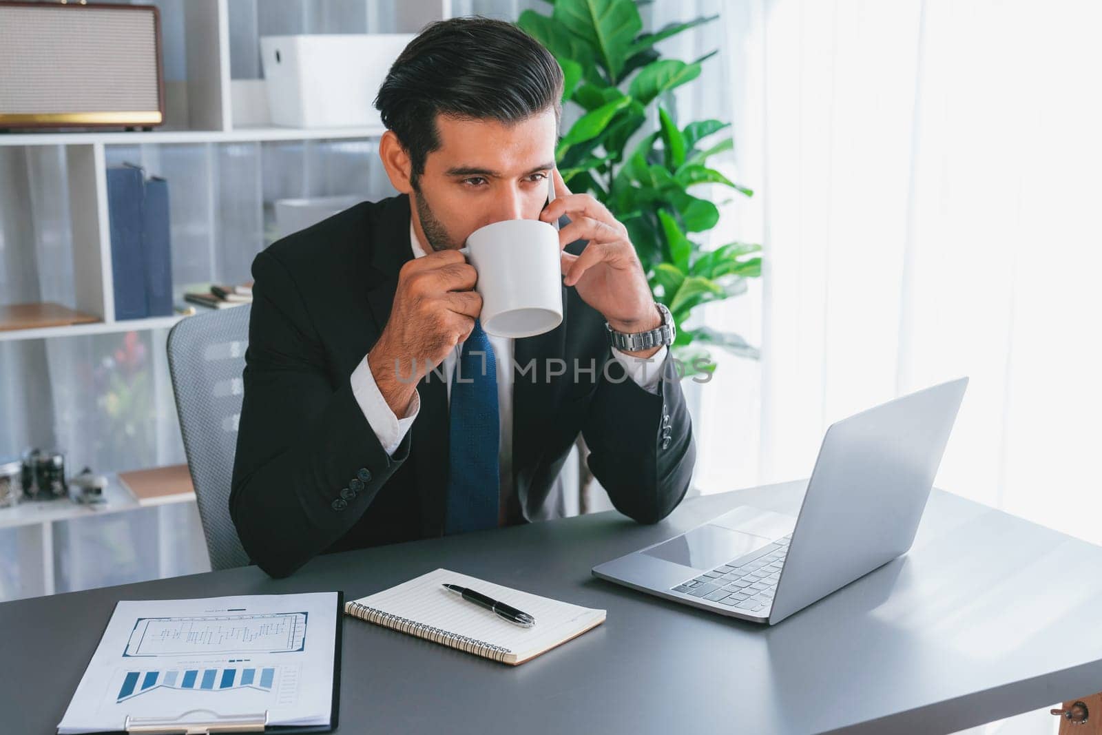 Businessman working in modern office workspace with cup of coffee in his hand while answering phone call making sales calls or managing employee. Fervent