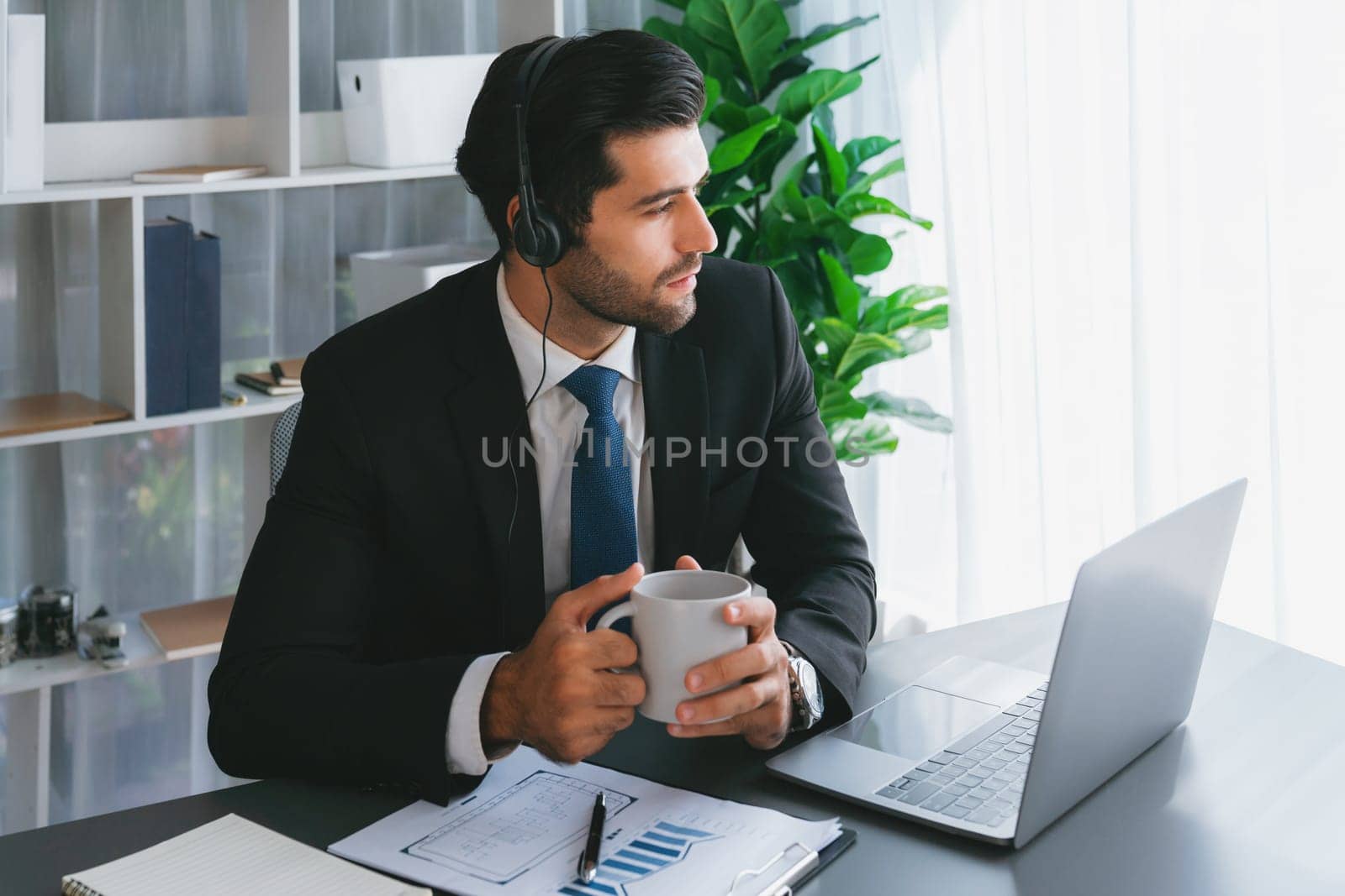 Manager of call center operator office sitting on his desk with his coffee while working on laptop. Office worker wearing headset and black suit working on customer support or telemarketing. fervent