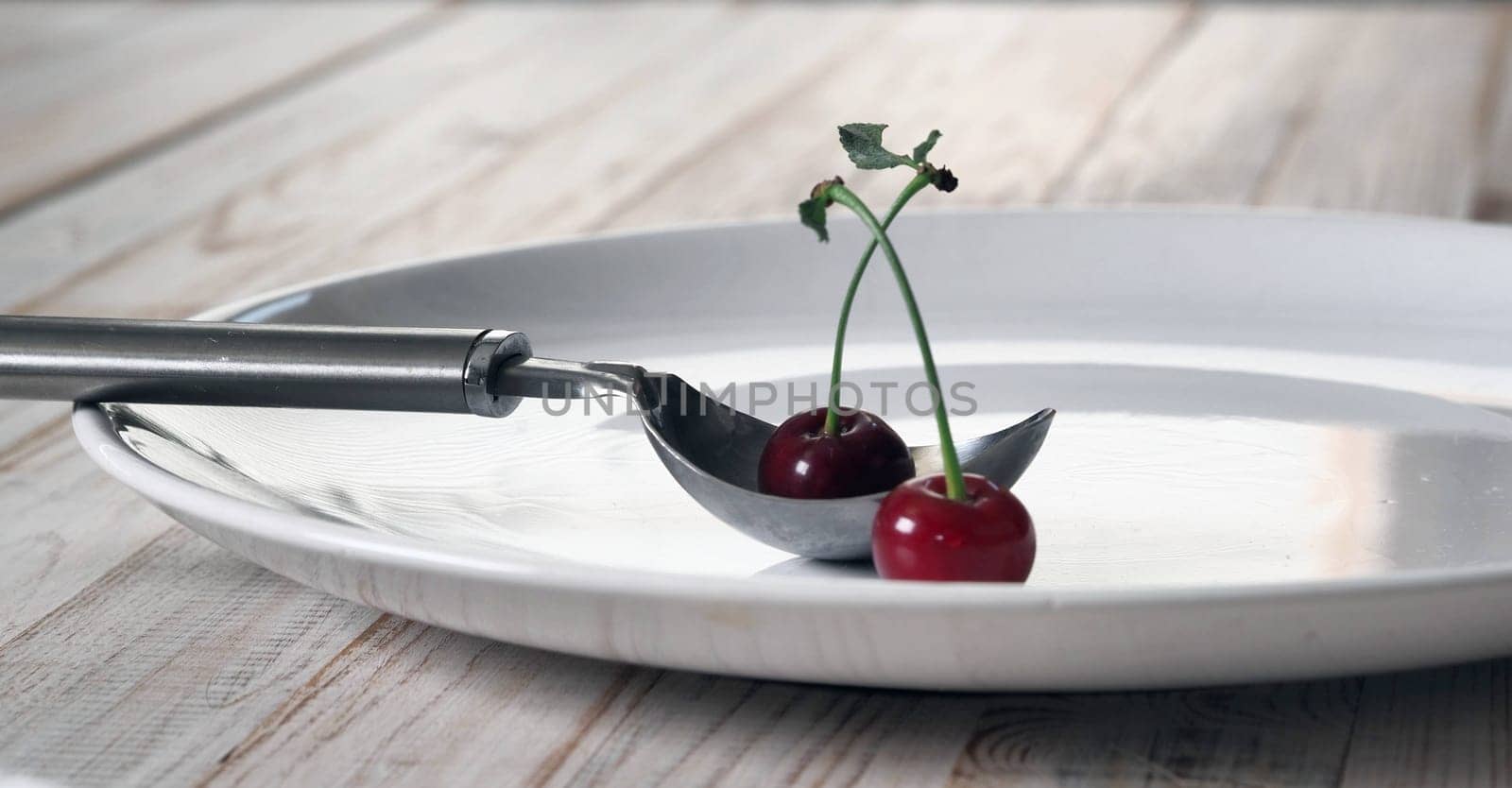 Red cherries in a white plate with a spoon for laying out ice cream on a white wooden table.Food background. by TatianaPink