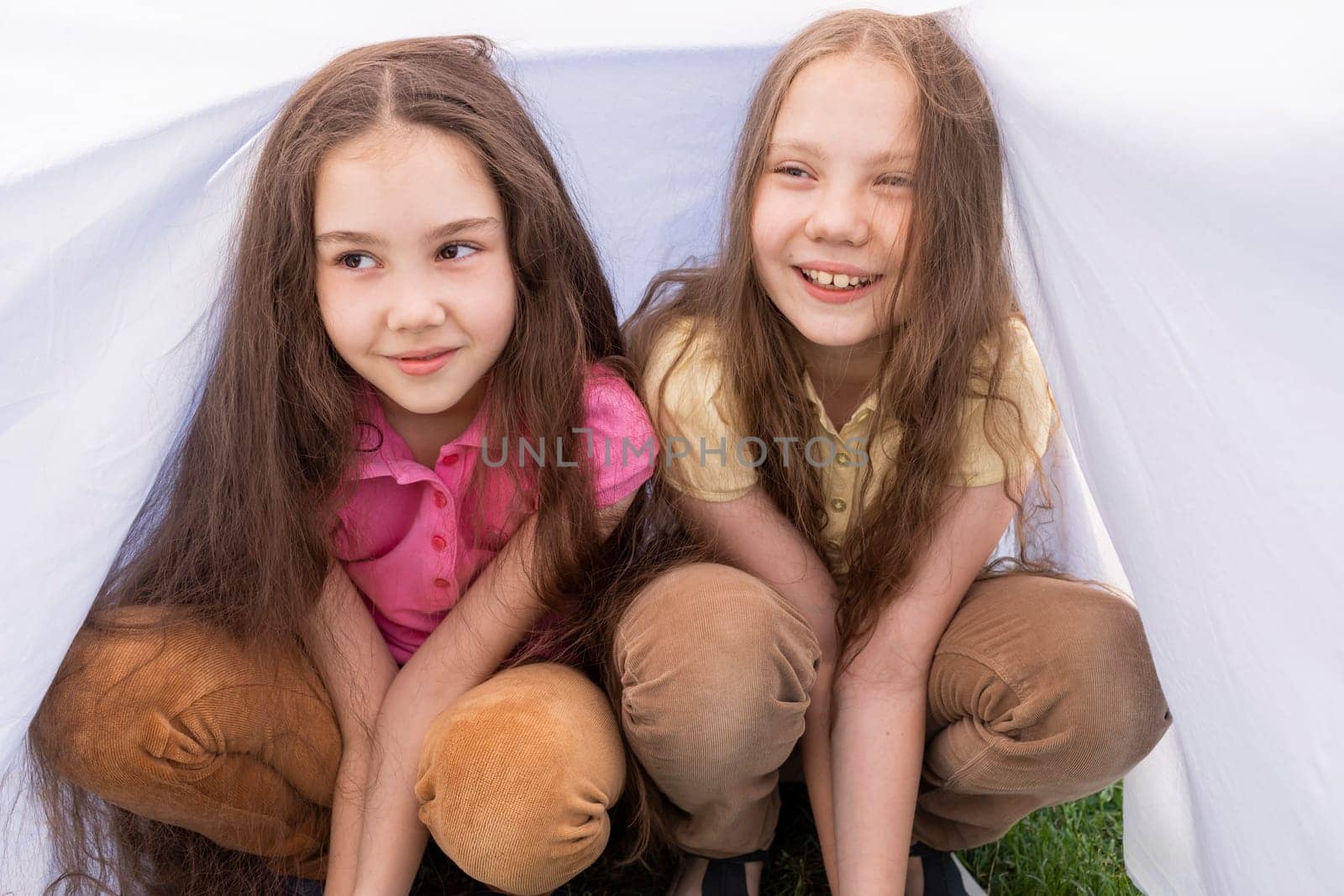 Two Cute Little Sisters With Long Hair Sits Under White Sheet On Grass in Meadow. Caucasian Asian Siblings Have Fun, Joy Together. Full Family, True Friendship. Horizontal Plane. High quality photo