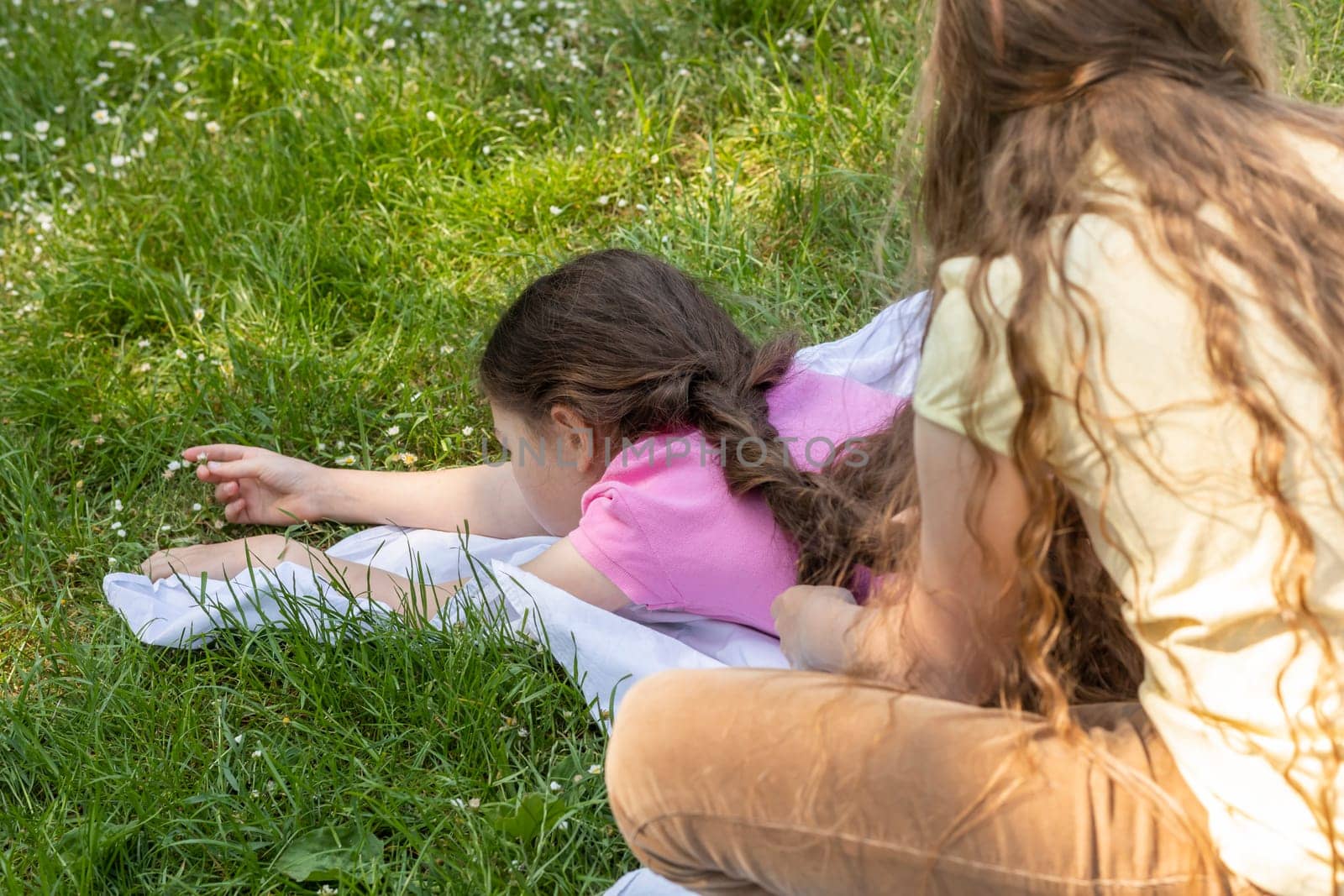 View From Back Little Girl With Long Hair braids Sister's Hair Sitting On Grass in Meadow At Sunny Day. Siblings Love, Carefree Time, True Friendship, Family Lifestyle. School Break. Horizontal Plane