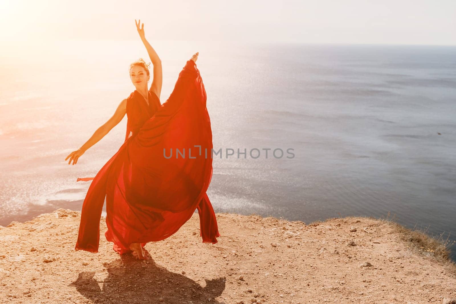 Side view a Young beautiful sensual woman in a red long dress posing on a rock high above the sea during sunrise. Girl on the nature on blue sky background. Fashion photo.