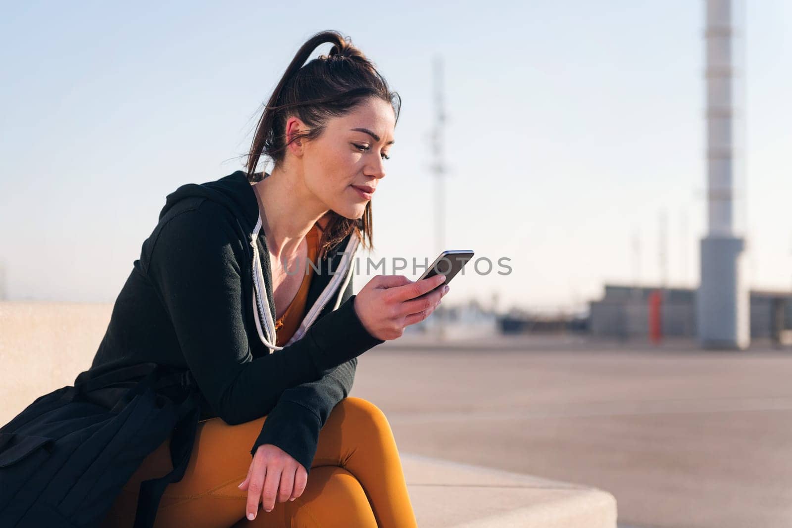 sportswoman sitting outdoors in the city using a mobile phone, concept of technology of communication and urban lifestyle, copy space for text