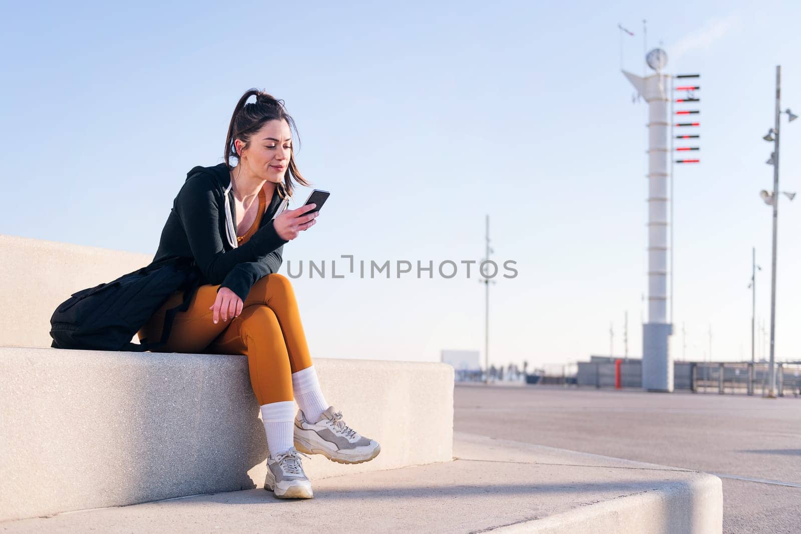 young sportswoman sitting outdoors in the city using a mobile phone, concept of technology of communication and urban lifestyle, copy space for text