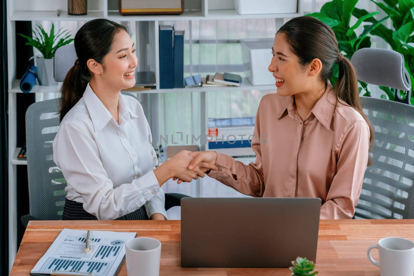 Two Asian businesswomen shaking hands in modern office after successfully working or analyzing using laptop as importance of teamwork and integrity in the workplace concept. Enthusiastic