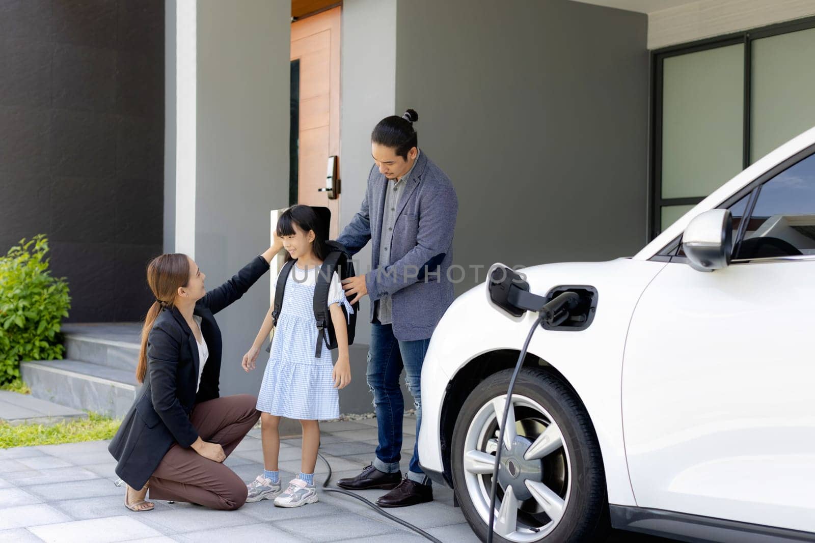 Progressive young parents and daughter with electric vehicle and home charging station. Green and clean energy from electric vehicles for healthy environment. Eco power from renewable source at home.