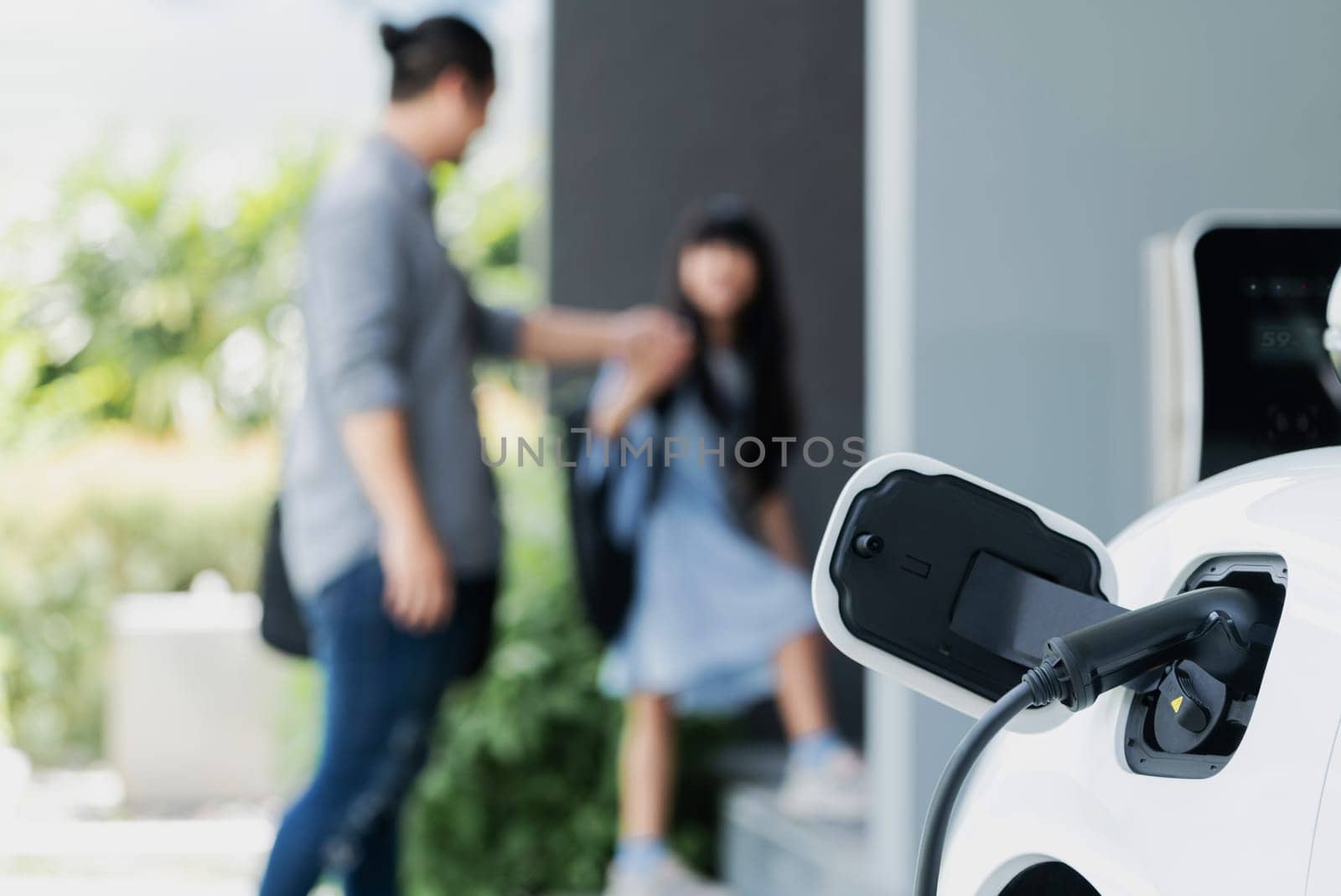 Focus electric car recharging at home charging station with blurred father and daughter walking in background. Progressive green and clean energy vehicle for healthy environment lifestyle concept.