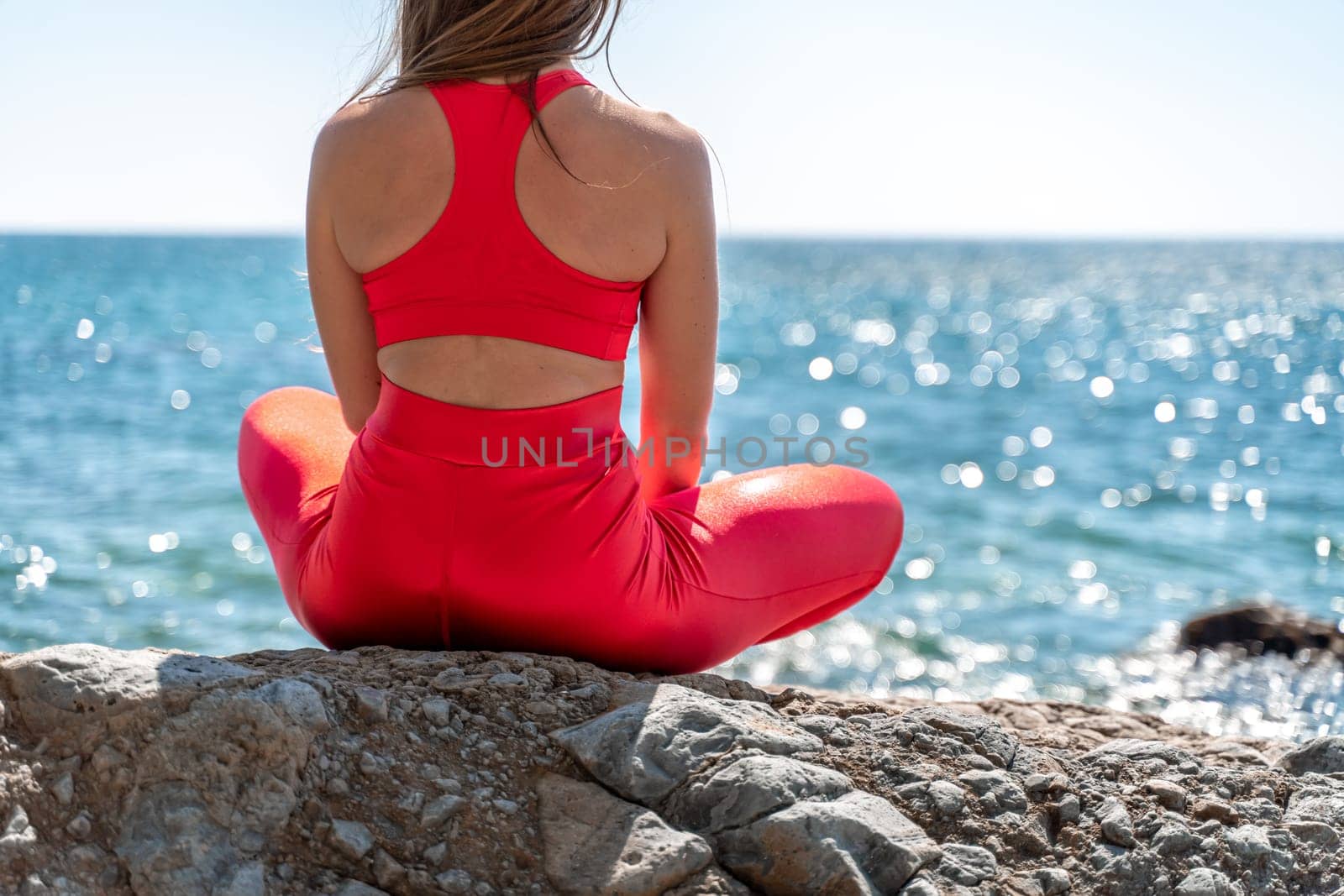 A young woman in red leggings and a red top with long loose hair practices yoga outdoors by the sea on a sunny day. Women's yoga, fitness, Pilates. The concept of a healthy lifestyle, harmony. by Matiunina