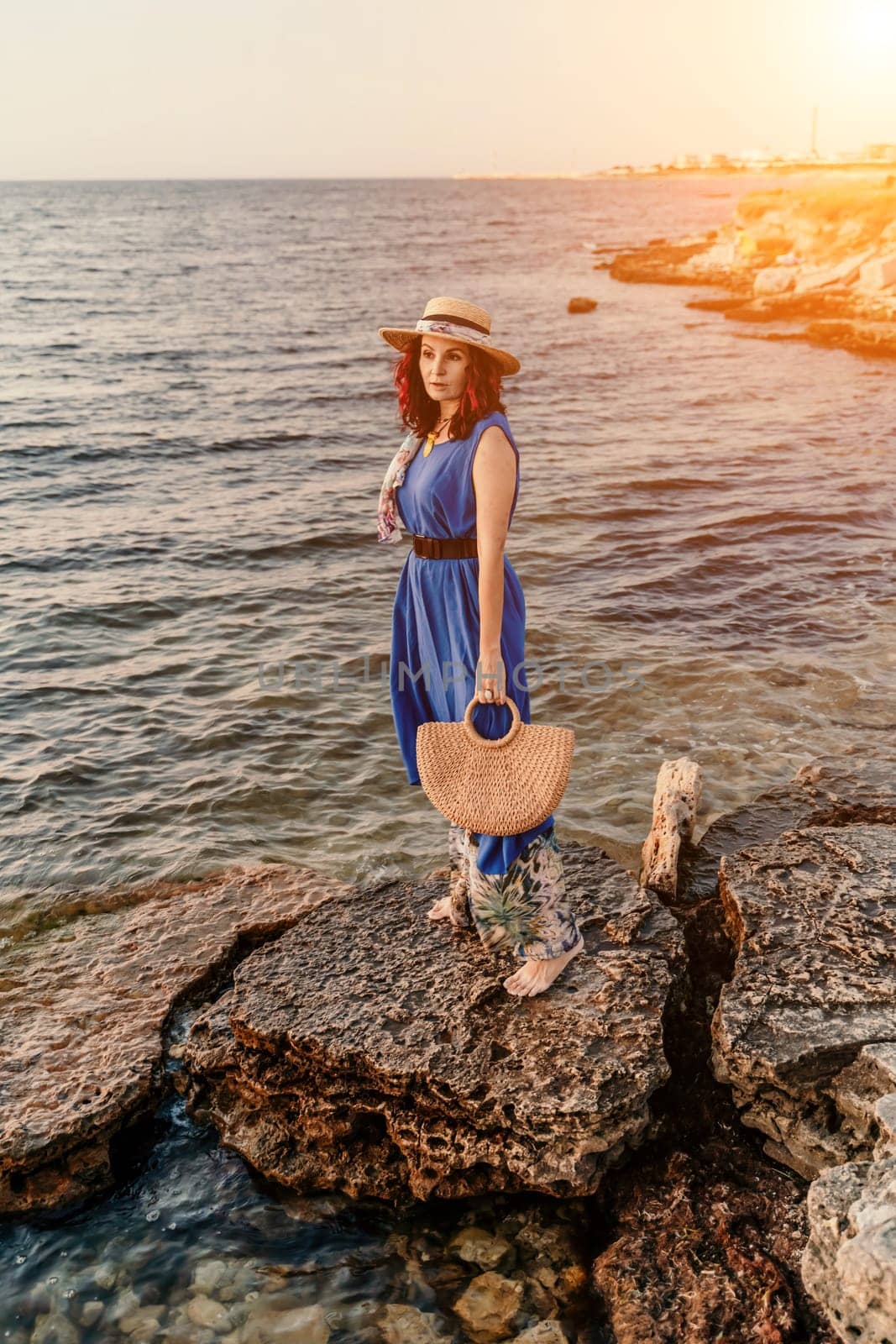 A woman in a dress, hat and with a straw bag is standing on the beach enjoying the sea. Happy summer holidays.