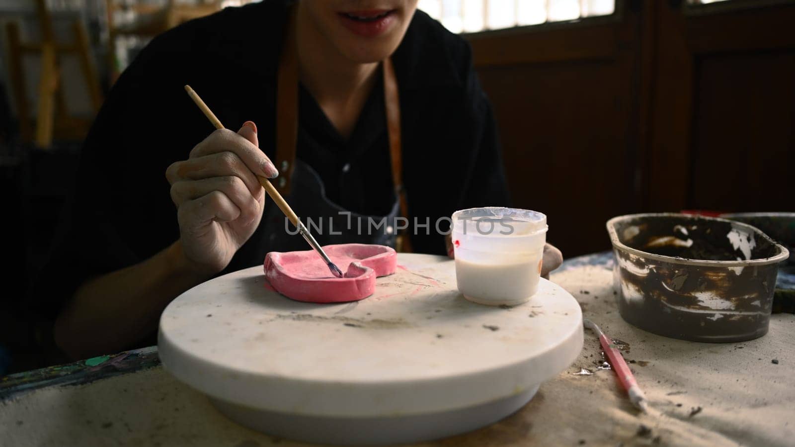 Cropped shot of man painting clay plate with a paintbrush at desk in ceramic workshop by prathanchorruangsak