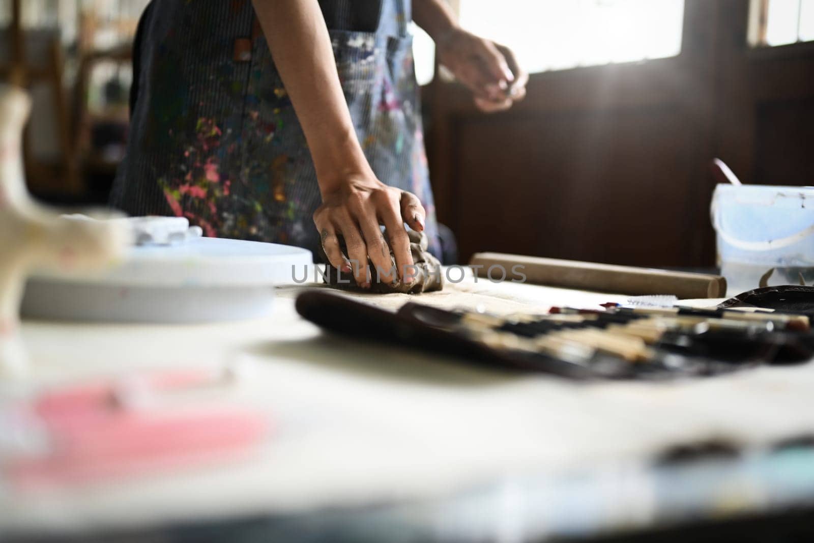 Craftswoman in apron kneading piece of raw clay with his hands on wooden table. Handicraft, creativity, hobby and activity concept by prathanchorruangsak