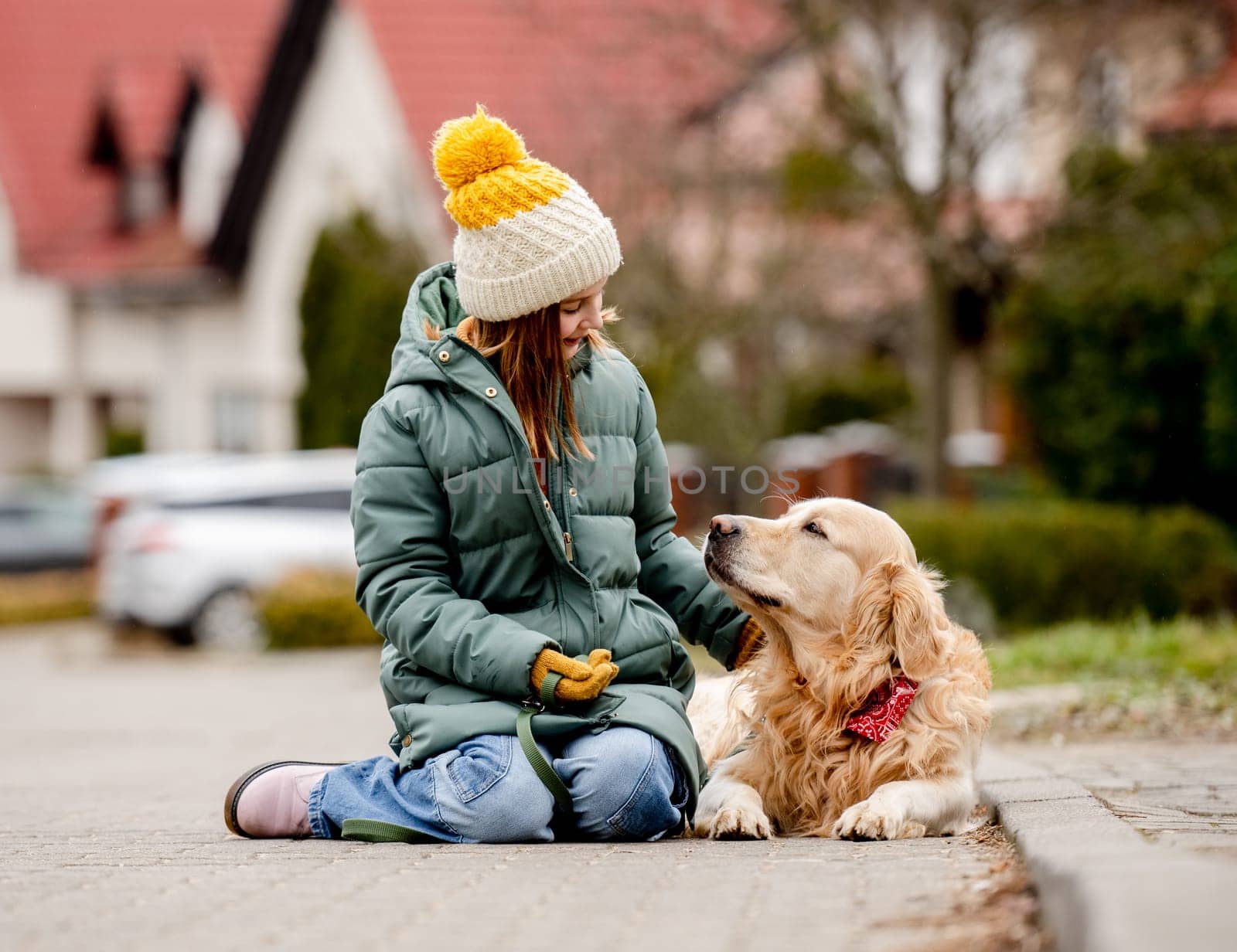 Preteen child girl sitting with golden retriever dog on asphalt at autumn street wearing hat and warm jacket. Pretty kid petting purebred pet doggy labrador outdoors at city