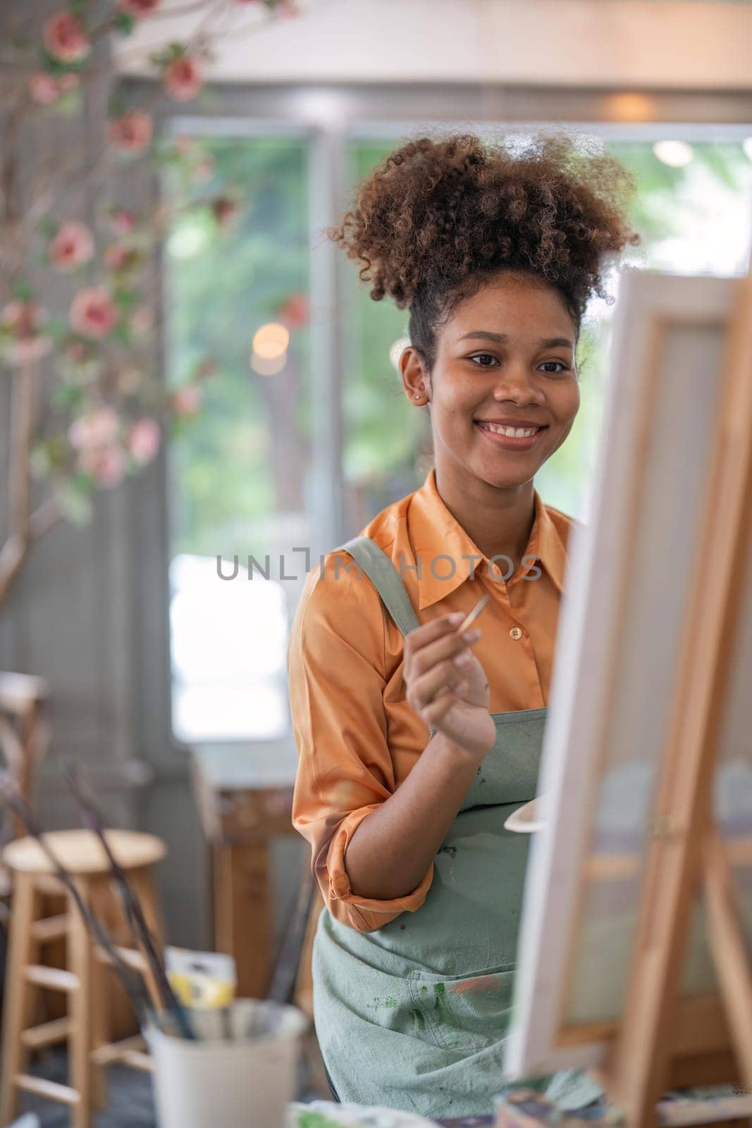 An African American woman who loves art or a female artist is holding a paint palette and paintbrushes while happily painting on paper at studio workshop.