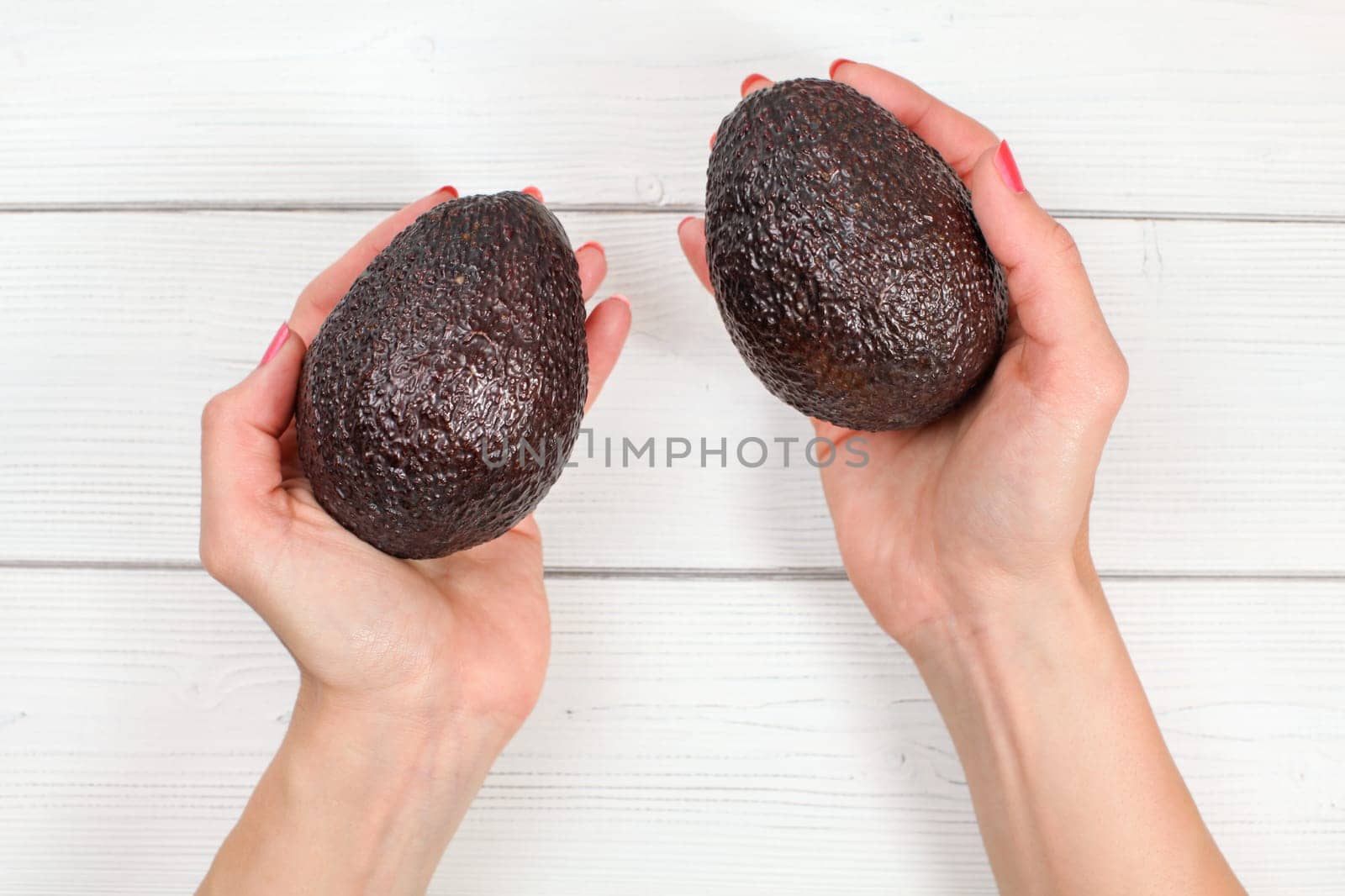 Looking down to woman hands holding two ripe brow avocados over white boards desk.