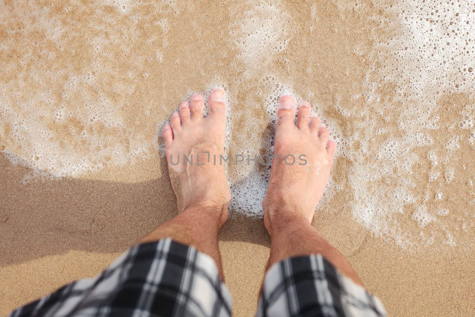 Looking down to man in shorts feet standing at wet beach sand, sea washing them, sun shining on.