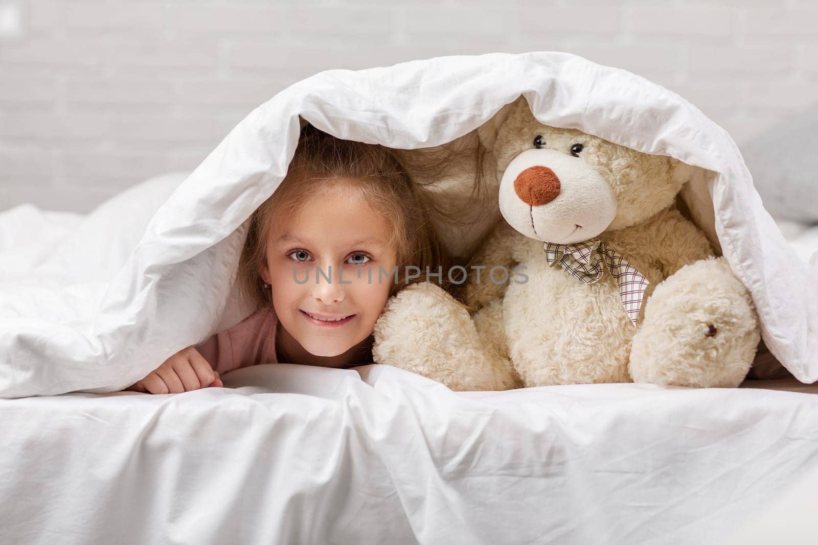 adorable little child girl playing with teddy bear in bed in morning. baby with a teddy bear under the covers