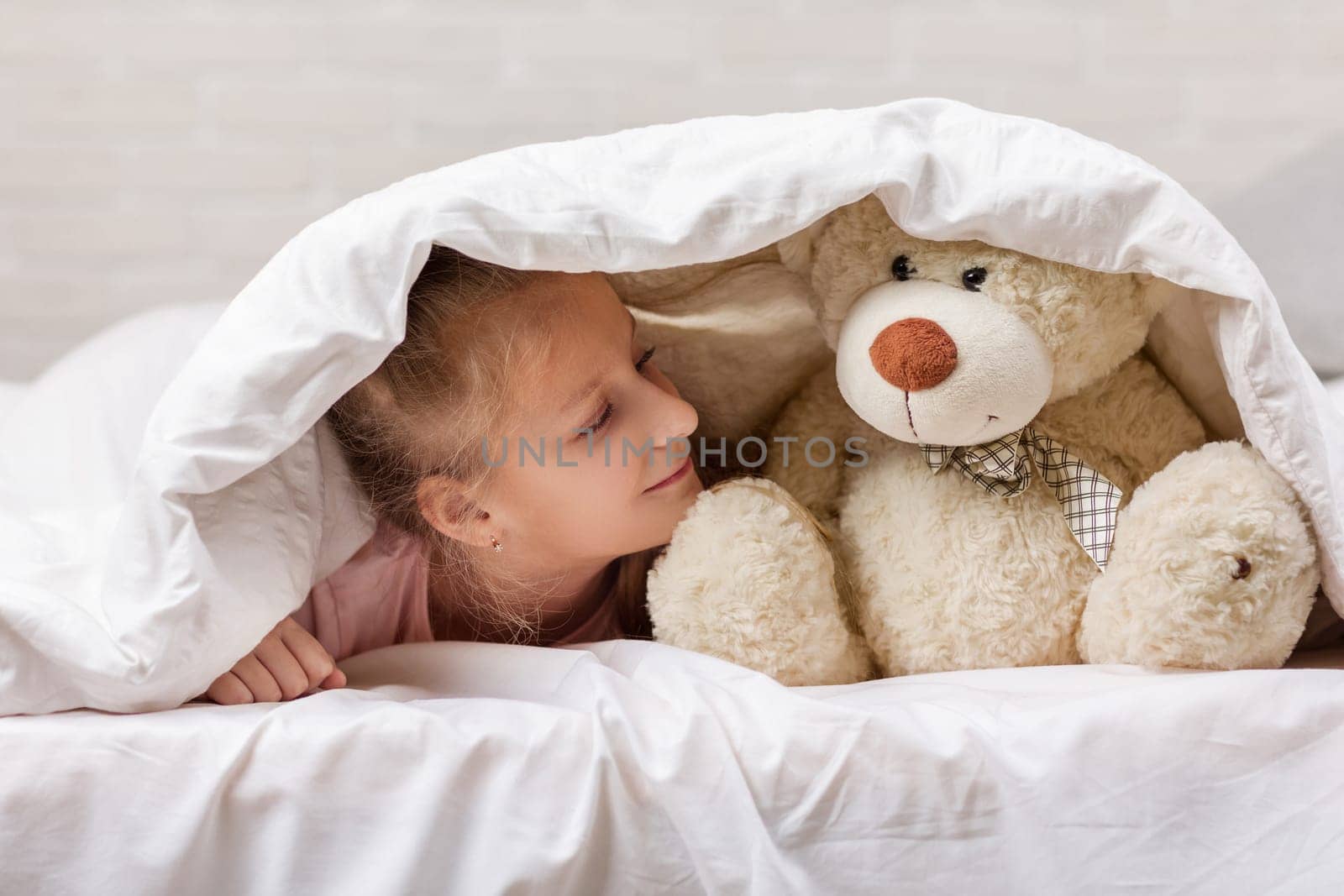 adorable little child girl playing with teddy bear in bed in morning. baby with a teddy bear under the covers