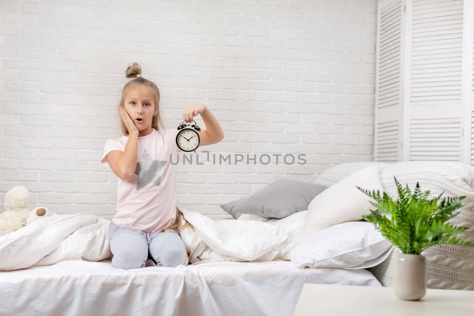 cute surprised little child girl in pyjamas with clock in bed. good morning