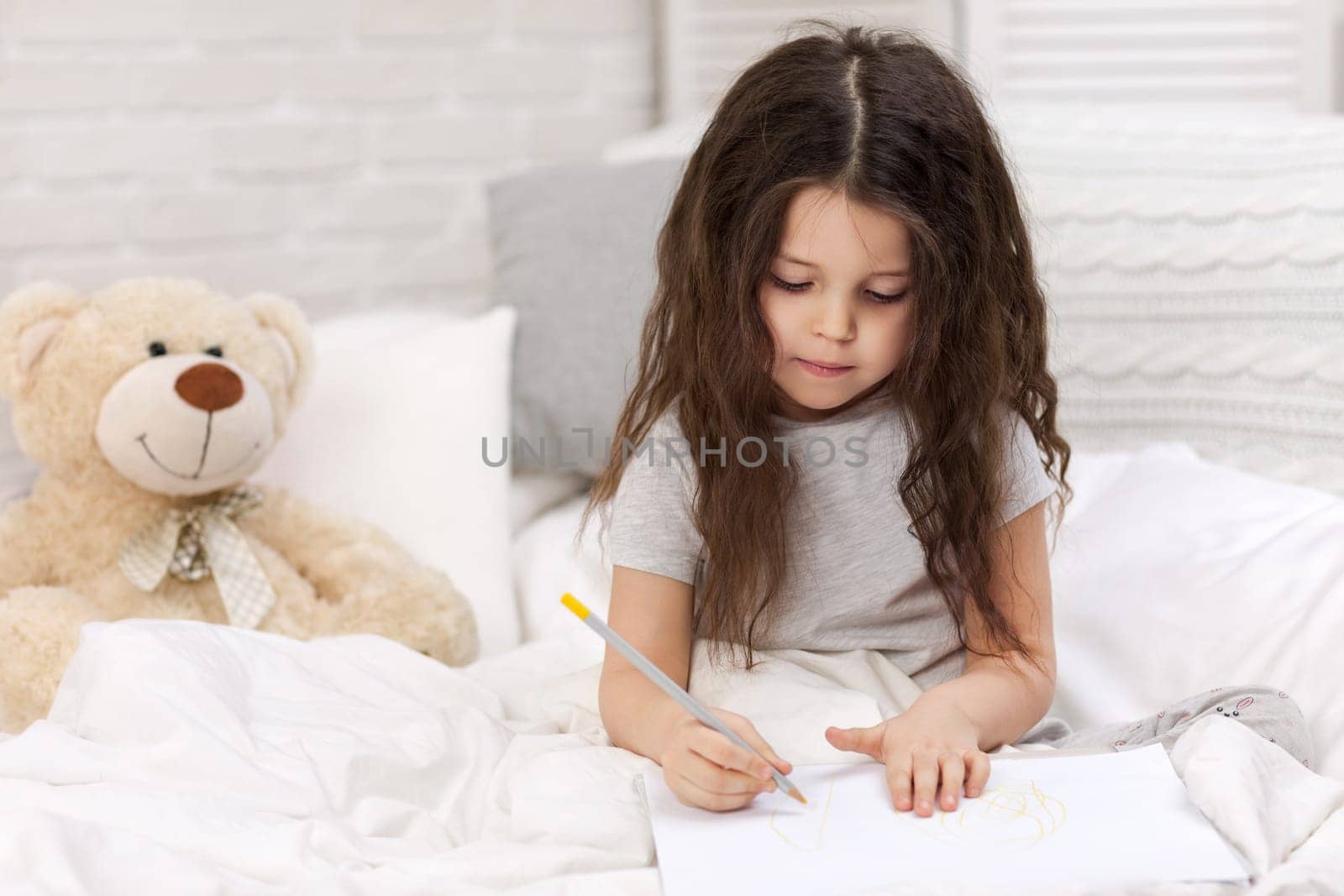 cute little girl with teddy bear drawing pictures while lying on bed. Kid painting at home