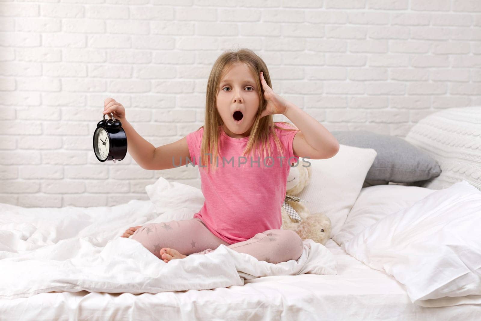 cute surprised little child girl in pyjamas with clock in bed. good morning