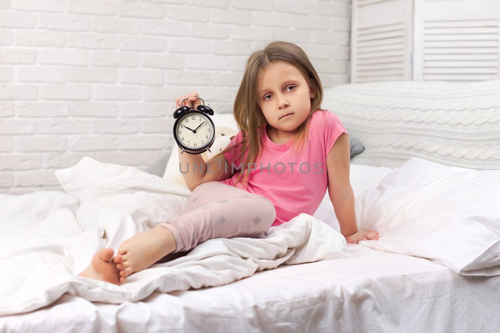 tired sleepy little child girl in pyjamas with clock in bed. good morning
