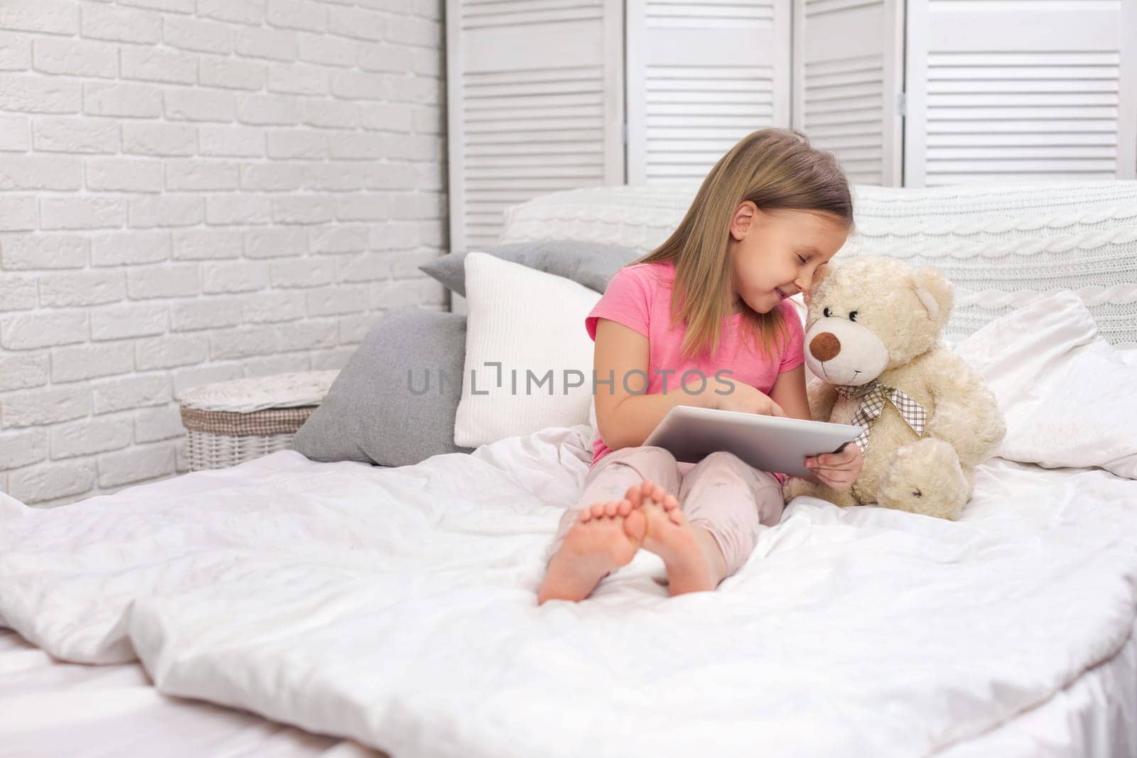 cute little child girl with teddy bear lies in bed uses digital tablet. child playing on tablet pc.