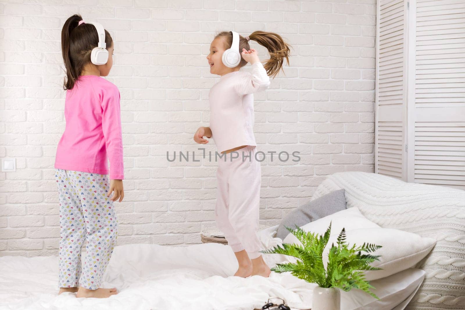 little children girls listening to the music with the headphones and dancing on bed. pajama party and friendship.