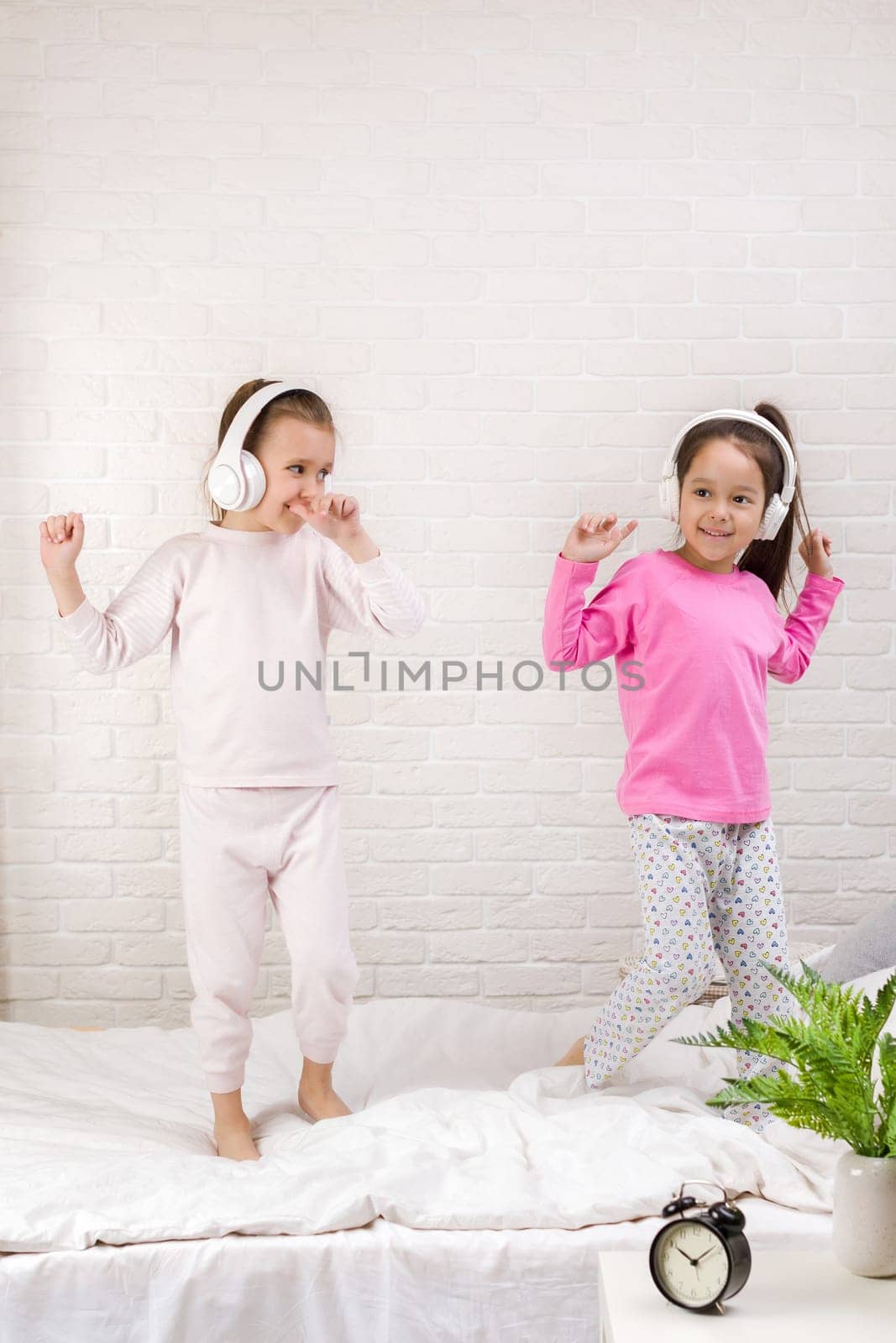 little children girls listening to the music with the headphones and dancing on bed. pajama party and friendship.