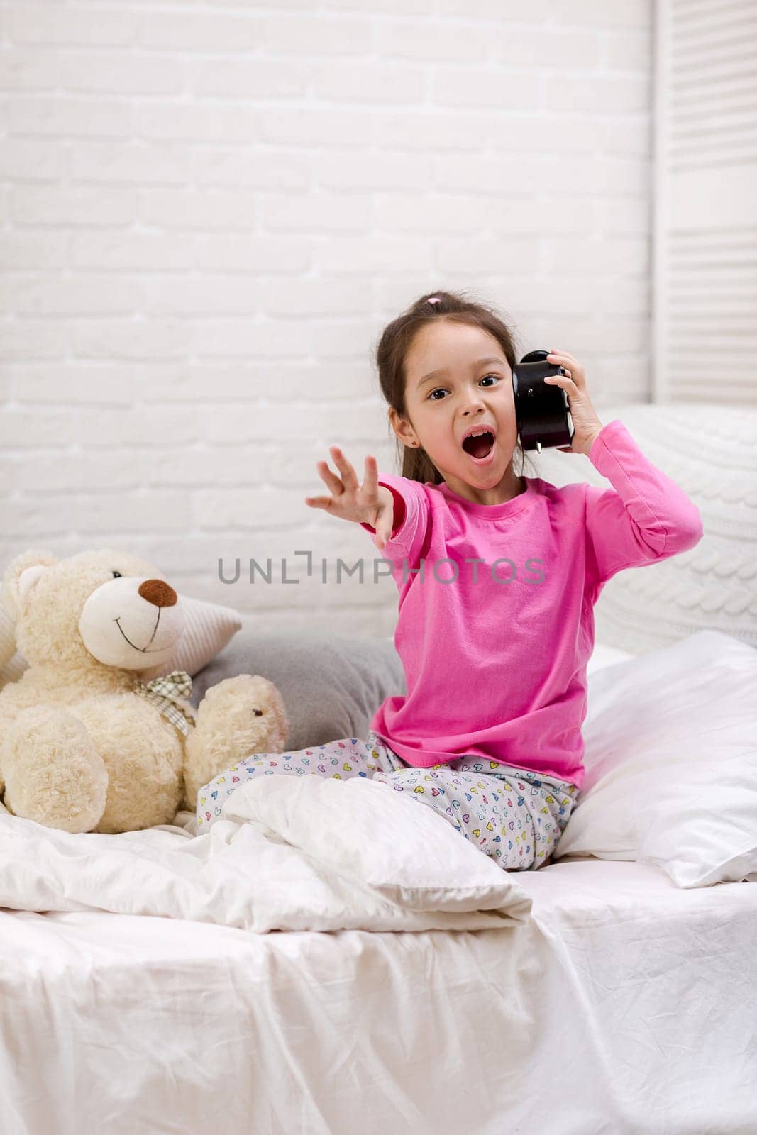 Surprised cute little child girl in pyjamas with clock in bed. good morning