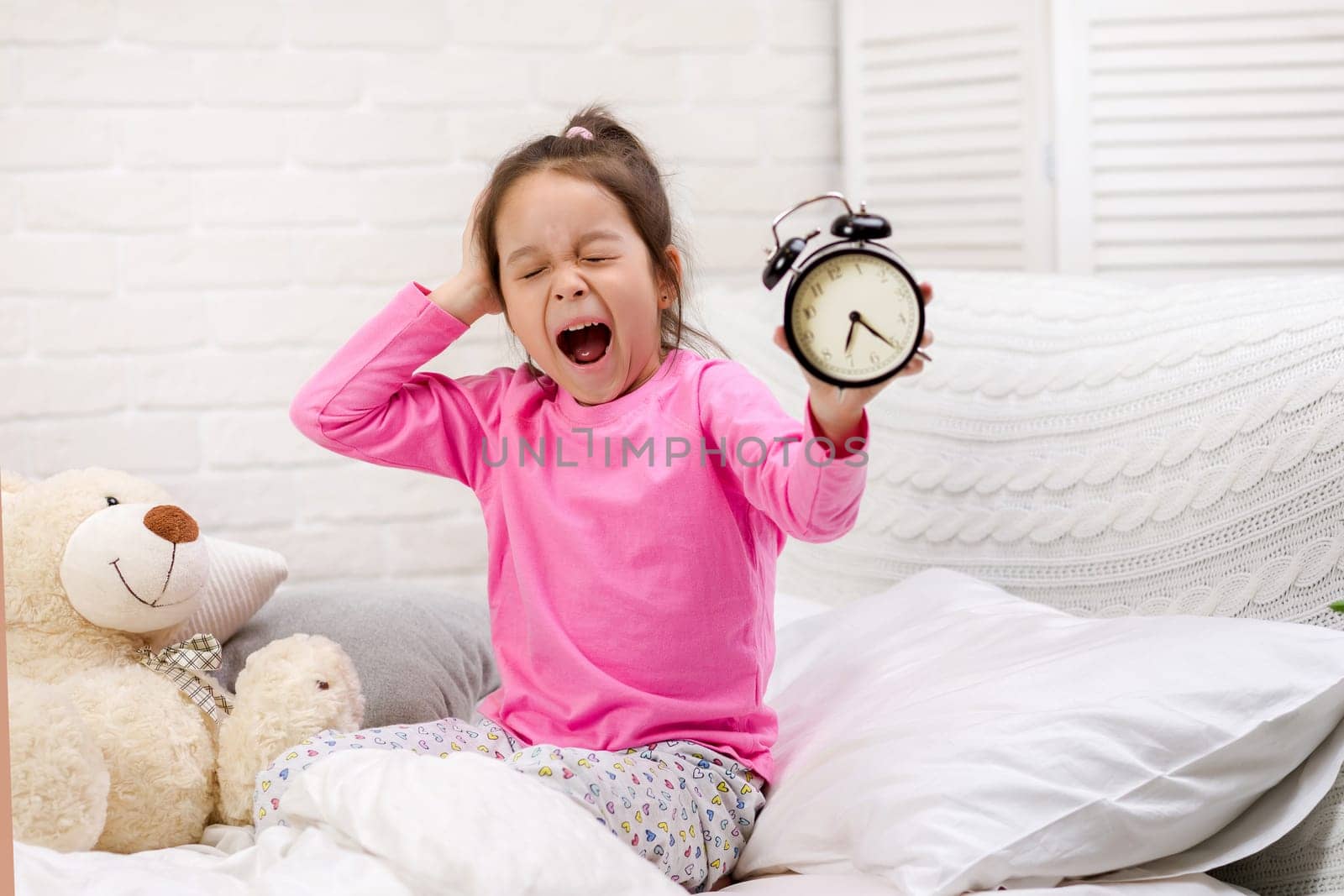 cute tired little child girl in pyjamas with clock in bed. good morning