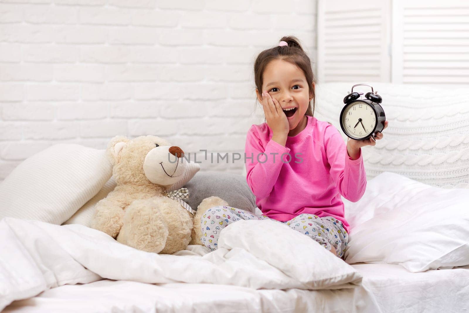 cute tired little child girl in pyjamas with clock in bed. good morning