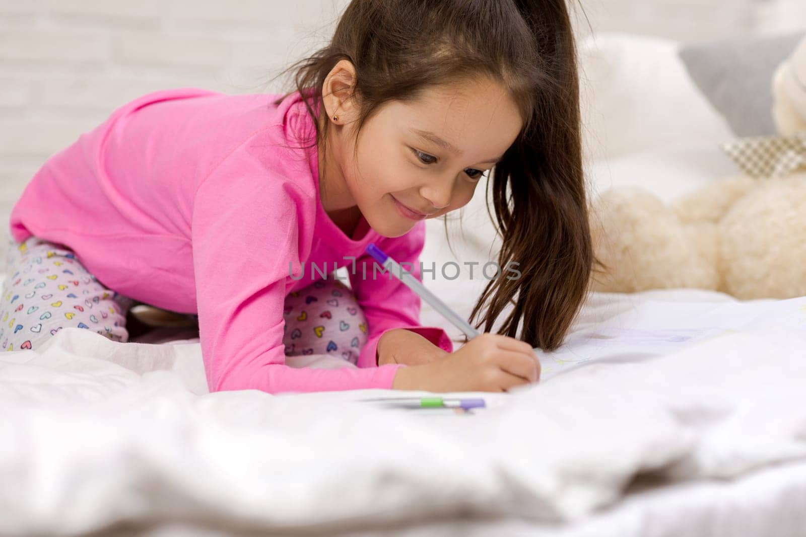 funny little girl drawing pictures while lying on bed. Kid painting at home