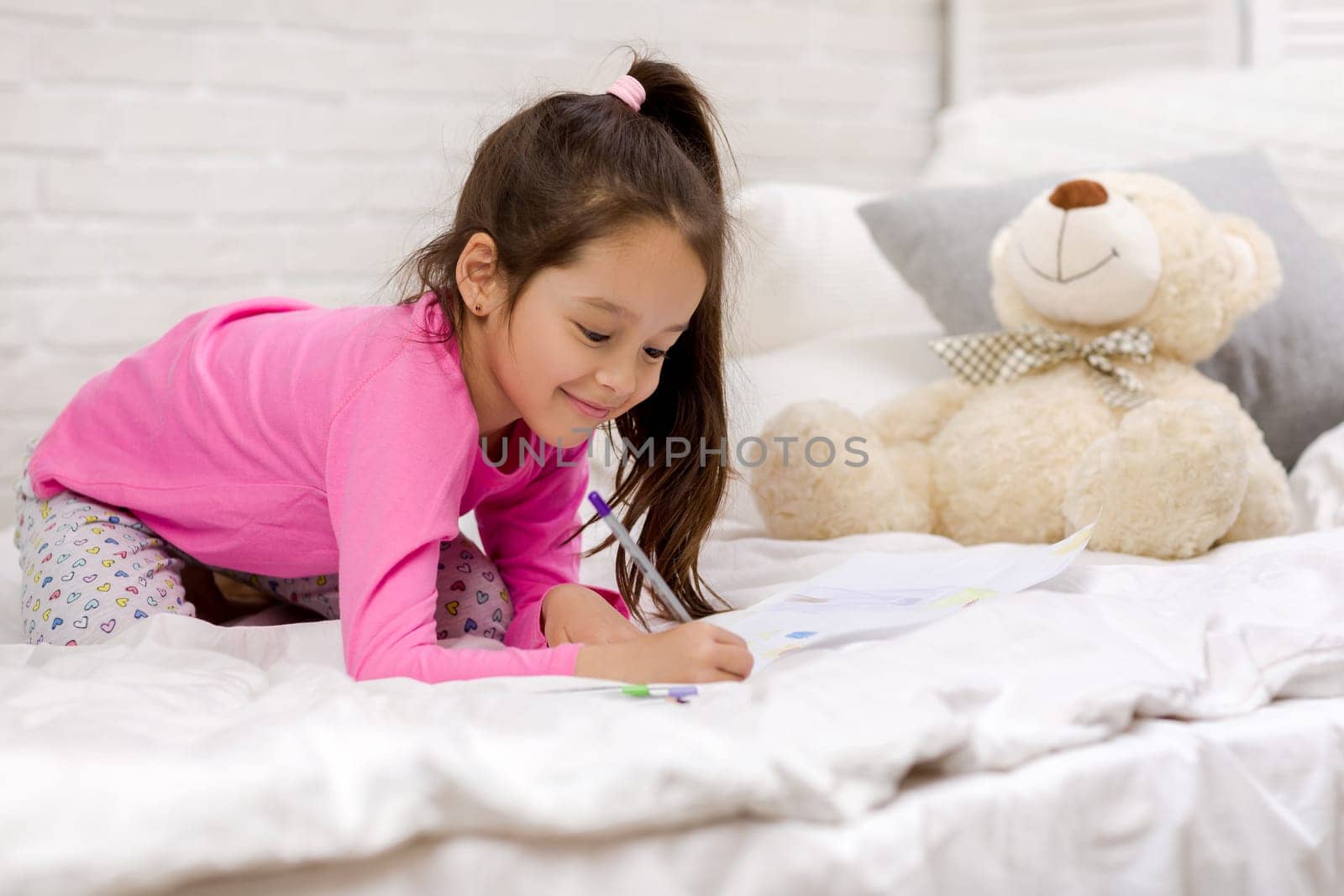 funny little girl drawing pictures while lying on bed. Kid painting at home