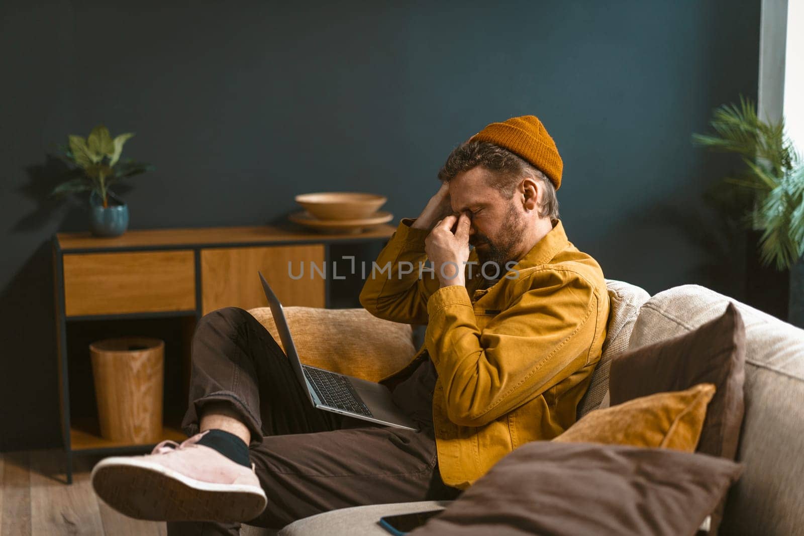 Sad and tired man is depicted sitting on a sofa at home. Overwhelmed by bad news or challenges, he holds his head with closed eyes in his hand, conveying a sense of emotional distress. by LipikStockMedia