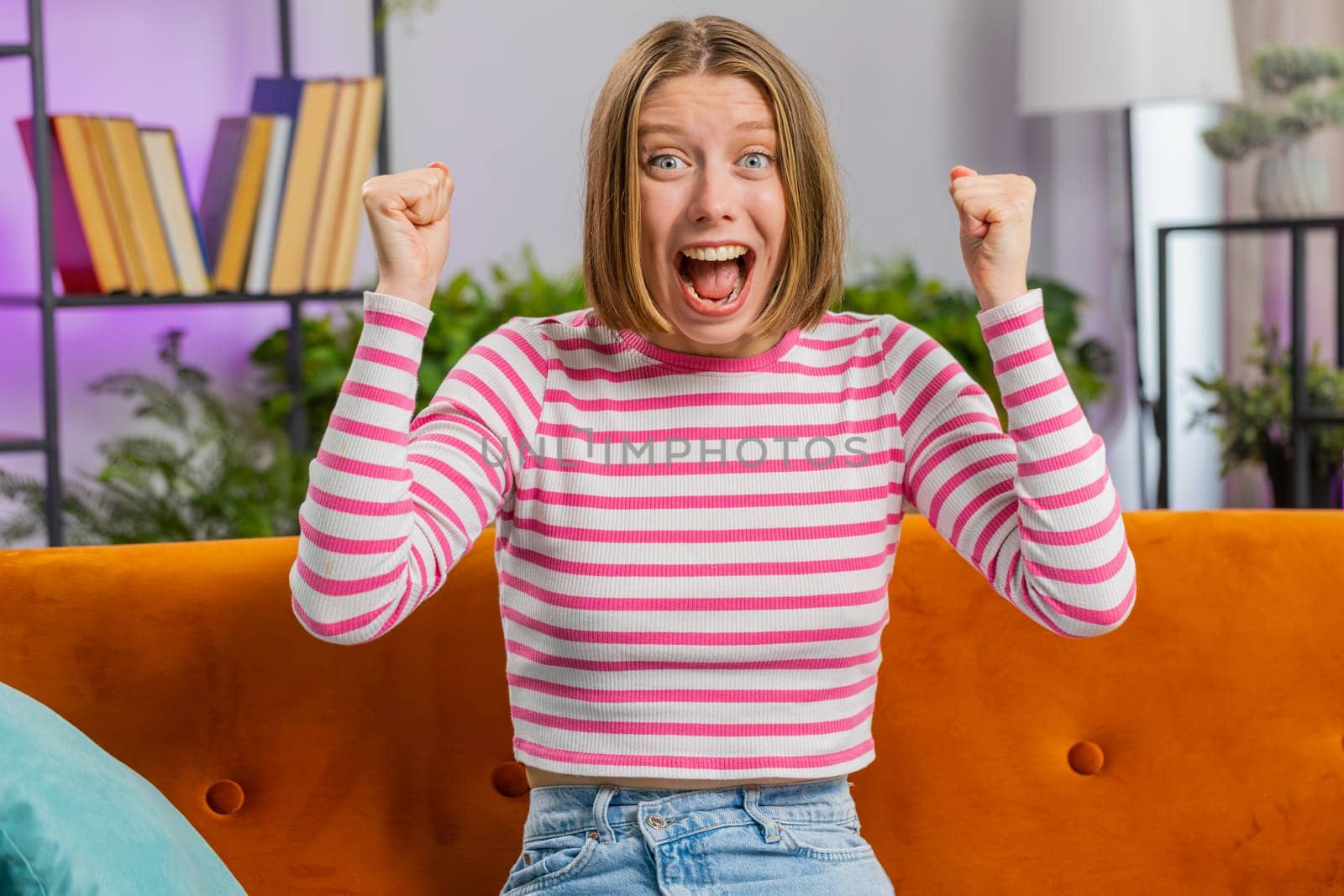 Happy young brunette woman shouting, celebrating success, winning, goal achievement good news raising fists in gesture I did it, lottery luck at home apartment. Girl in living room sitting on couch