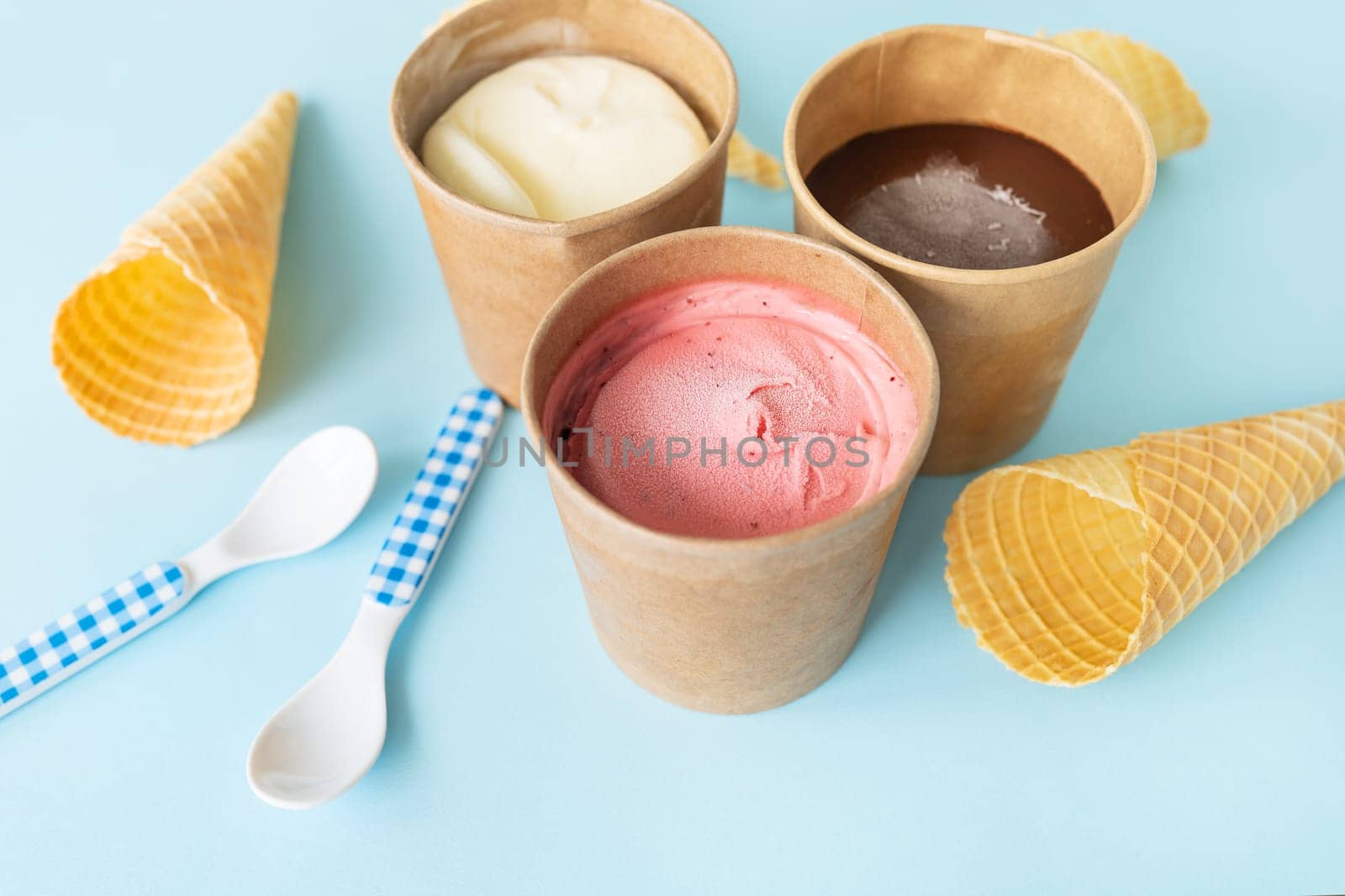 Ice cream in a craft box with different flavors - strawberry, chocolate, vanilla stands on a blue background along with a waffle cone and a checkered spoon. View from above