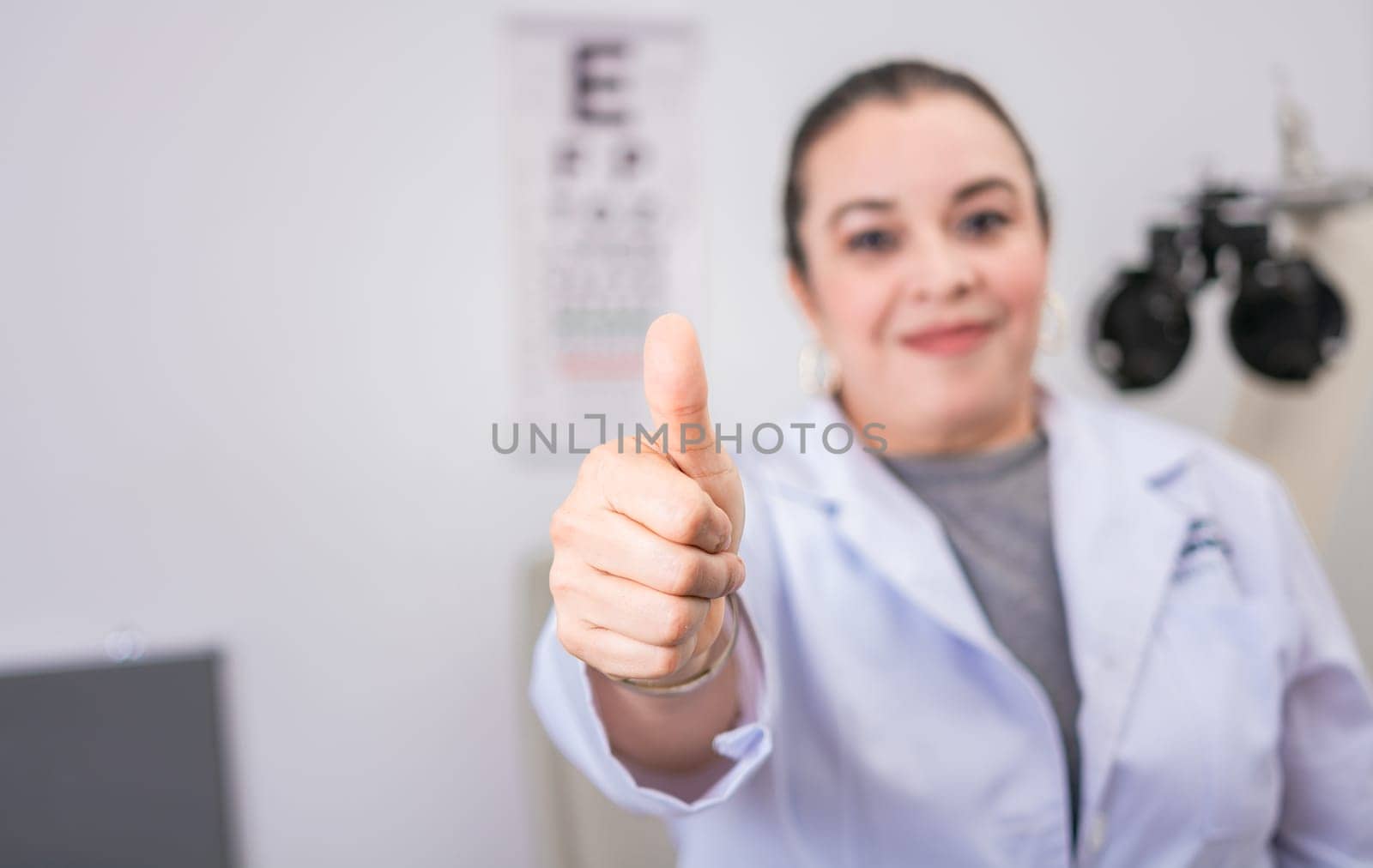 Female optometrist with thumb up in the laboratory. Optometry specialist with thumb up. Smiling eye doctor with thumb up in office by isaiphoto