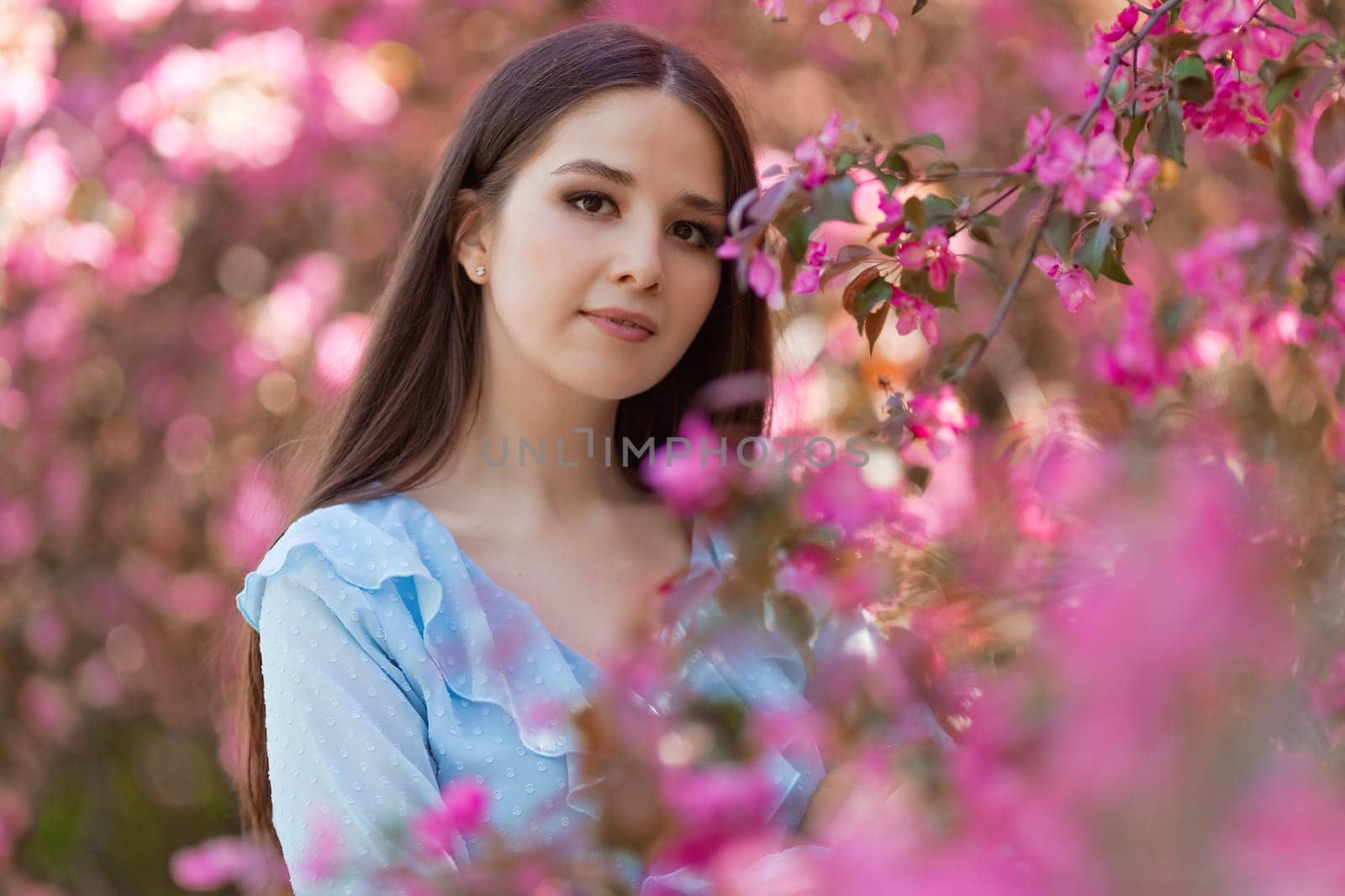 Portrait of adorable brunette girl with long hair is standing near a pink blooming apple tree, in the summer in the park. Close up