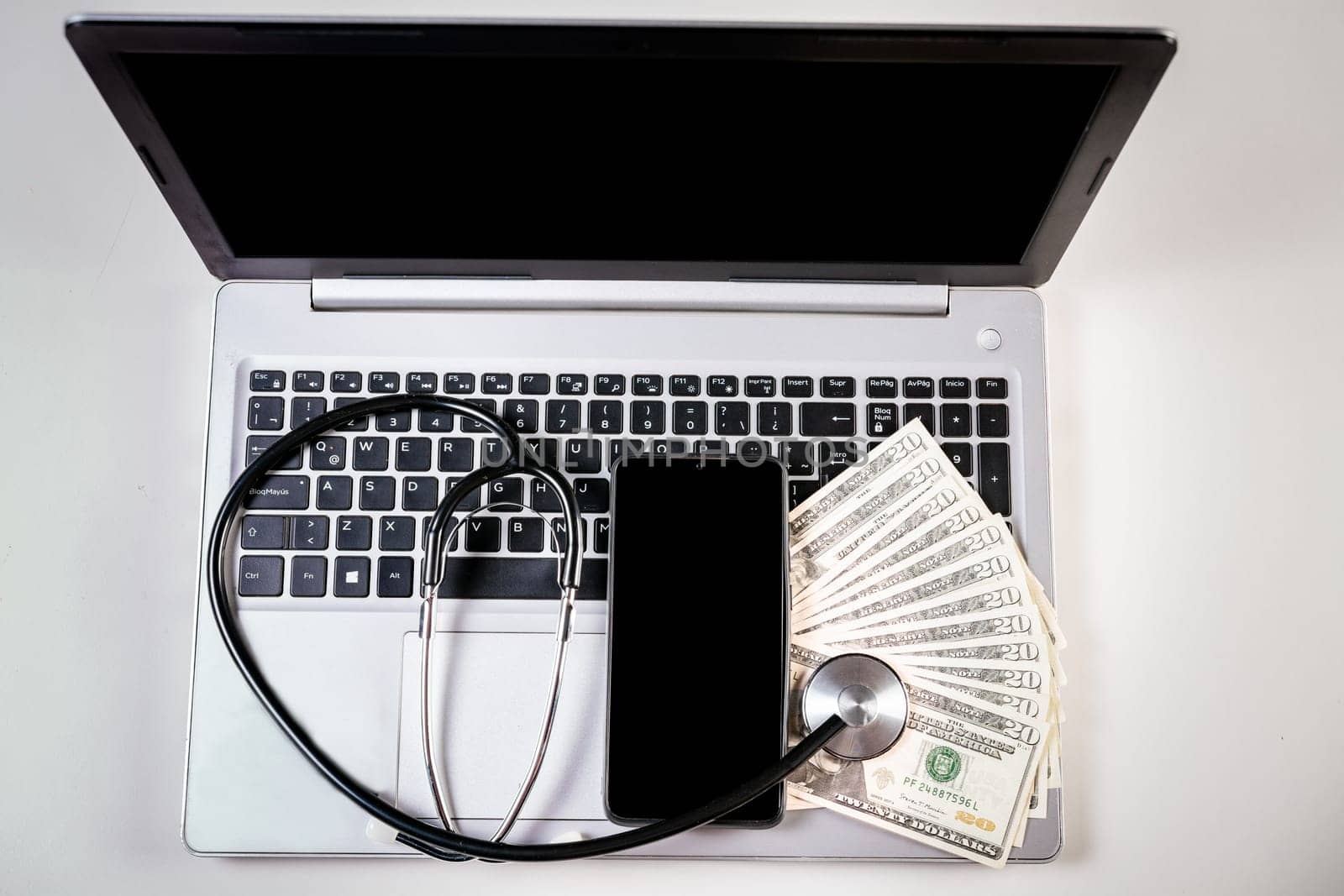 Mobile phone with dollar bills and stethoscope on laptop keyboard. Stethoscope on top of dollar bills on laptop keyboard by isaiphoto