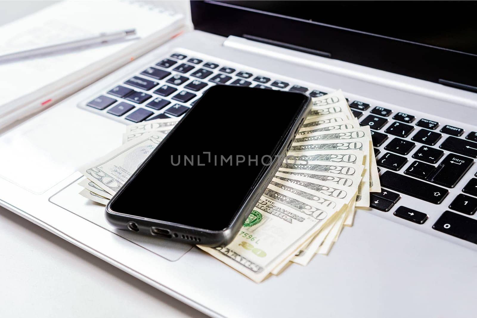Cell phone on laptop keyboard and dollar bills. Mobile phone on top of dollar bills on laptop keyboard. Top view of laptop with cell phone and dollar bills by isaiphoto