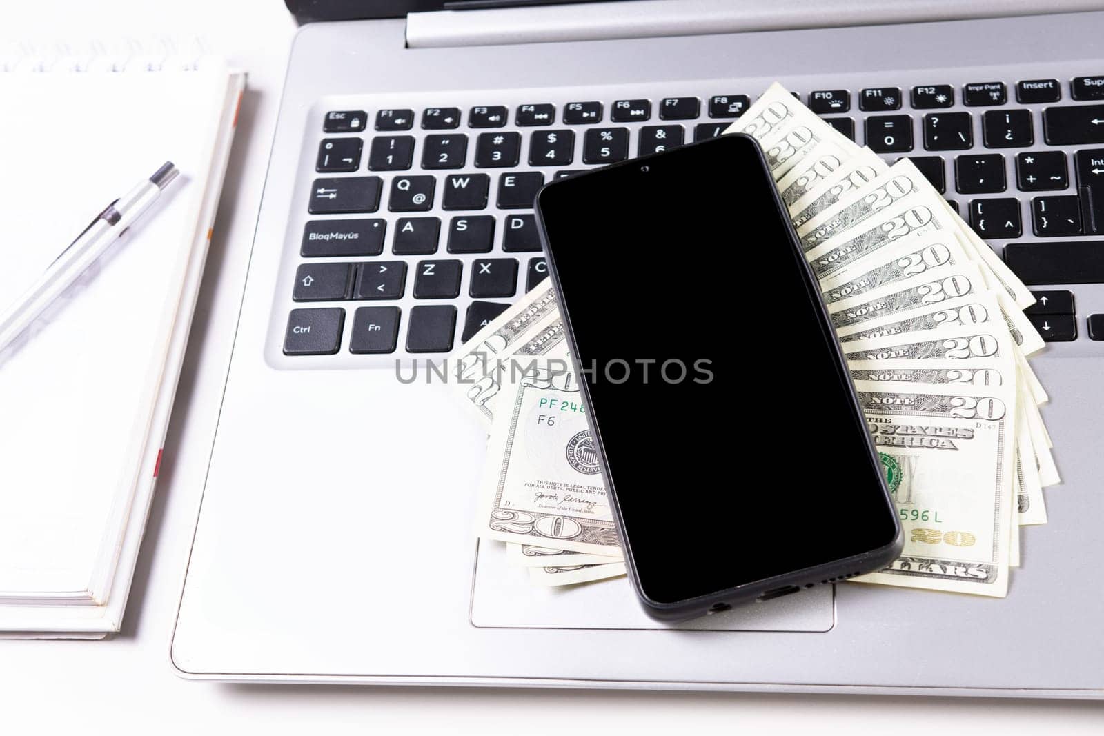 Mobile phone on top of dollar bills on laptop keyboard. Top view of laptop with cell phone and dollar bills. Cell phone on laptop keyboard and dollar bills by isaiphoto
