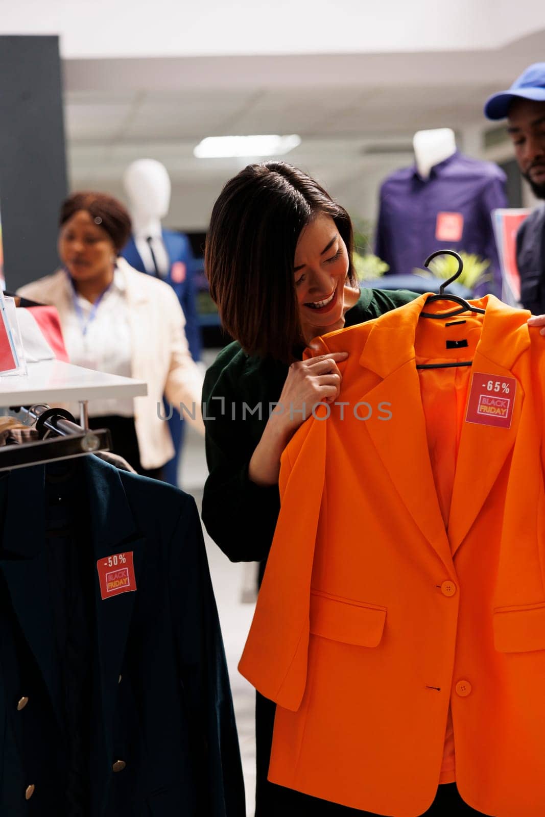 Satisfied Asian woman shopper trying on classic orange jacket with red price tag while shopping on Black Friday in clothing store. Smiling girl customer feeling happy while buying clothes on sale