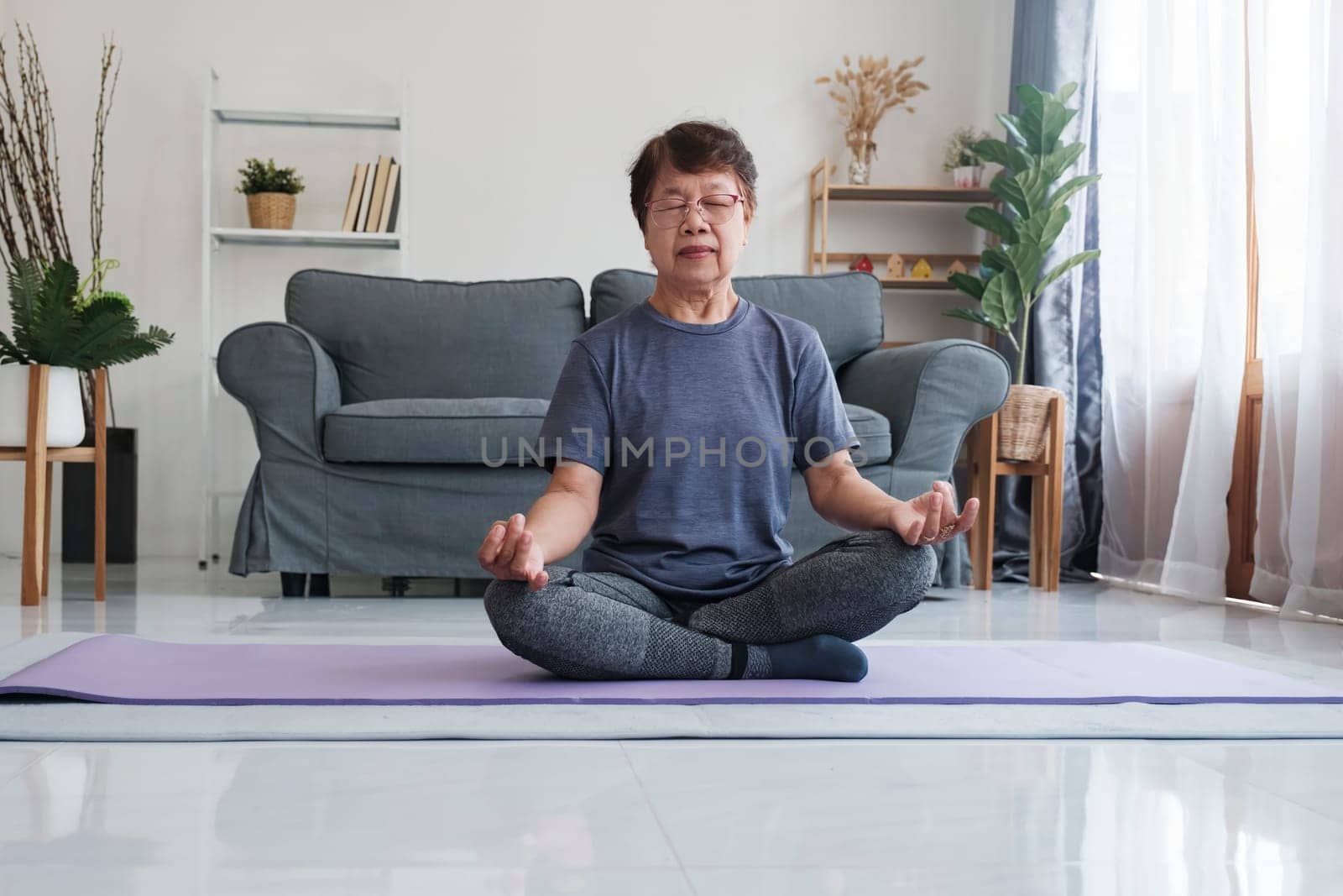 A senior woman finds inner peace through yoga and meditation at home, embracing relaxation and tranquility of the mind. Calm barefoot middle aged female sitting on carpet in half lotus posture, making mudra gesture and closing eyes, having peaceful facial expression, doing mindful meditation, concentrating on breathing.