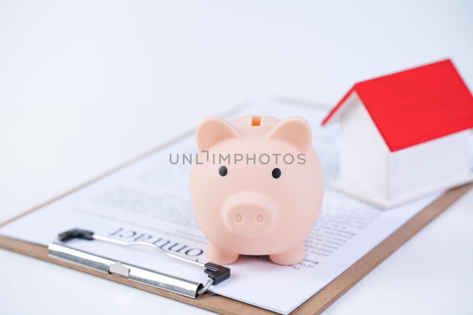 Piggy bank, beautiful wooden house model over a signed contract on white background, concept of saving money to buy a home insurance, close up, copy space. by ROMIXIMAGE