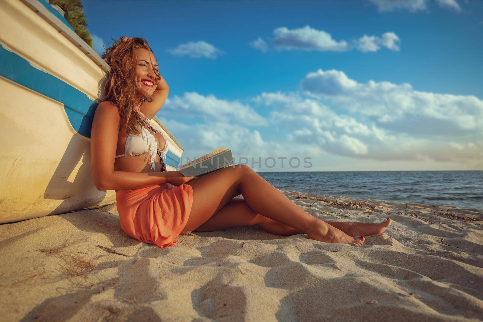 Young woman reading a book on the sandy beach.  She is sitting next to boat and enjoying with closed eyes and smile on her thinking face.